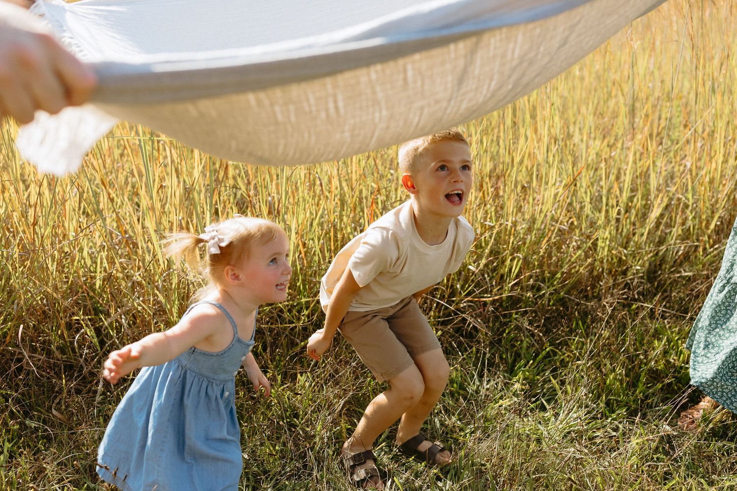 Two young children playing joyfully in a golden field under a white fabric canopy on a sunny day.