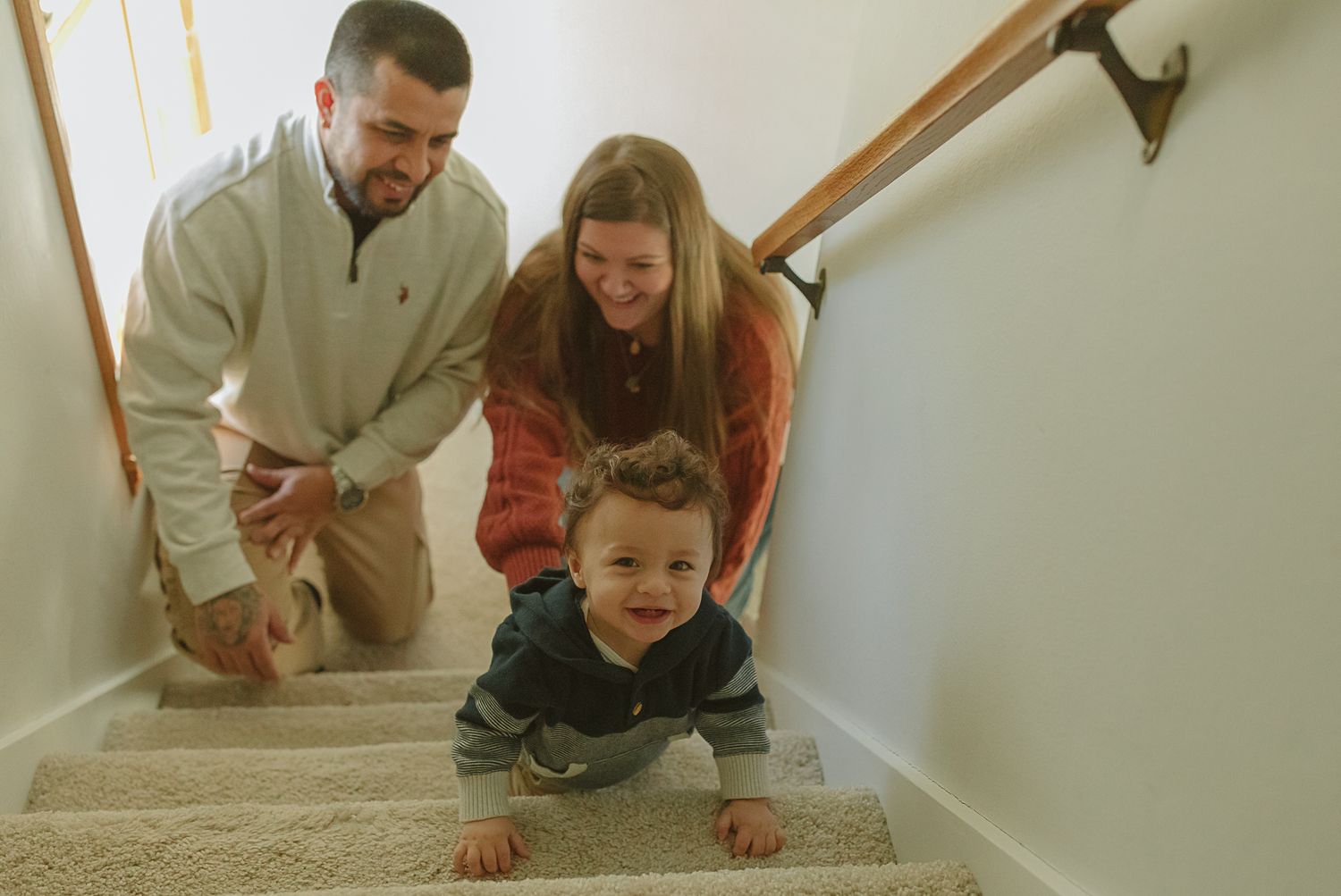Happy parents watching their toddler crawl up carpeted stairs inside their home.