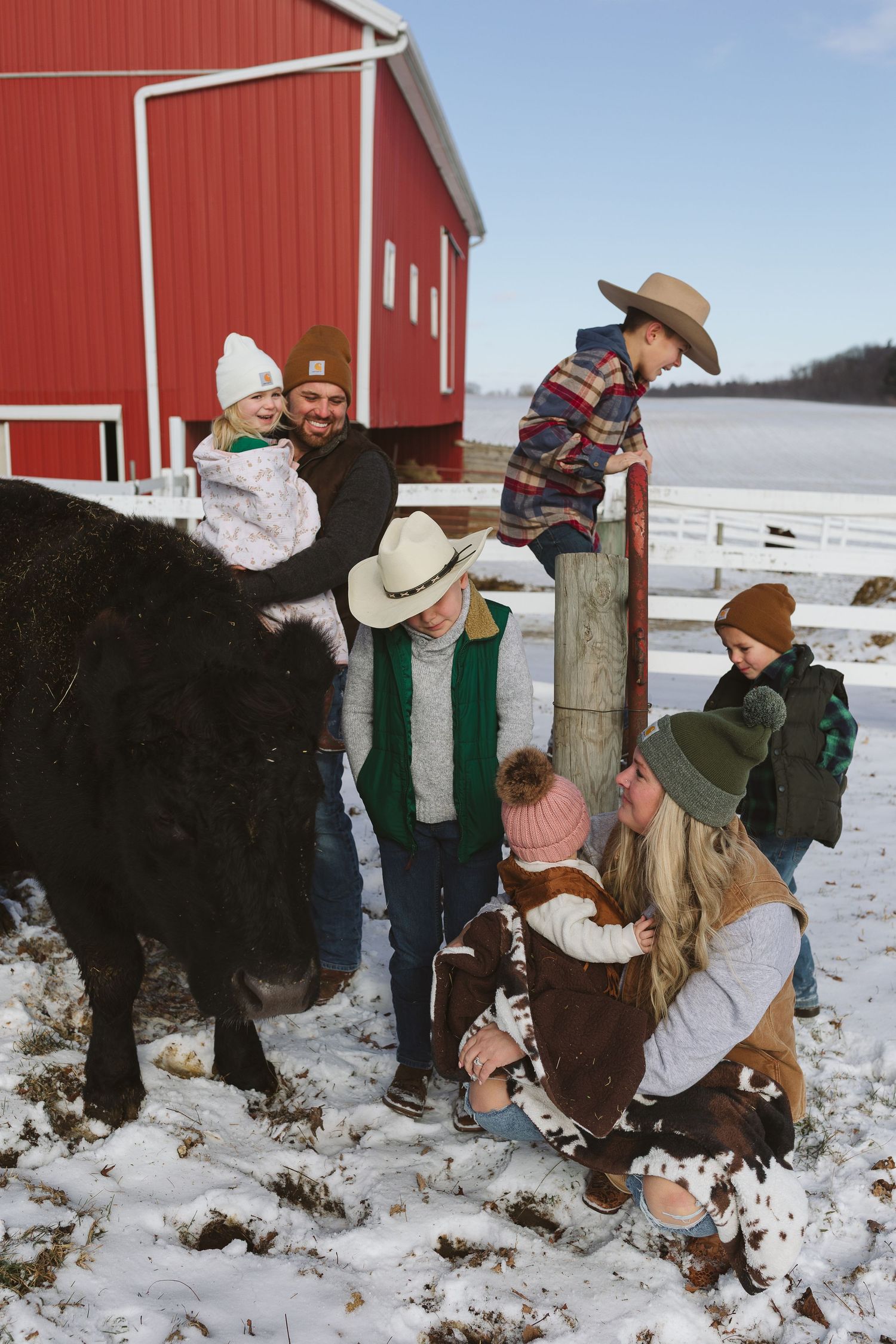 Family with children petting cattle outside red barn on snowy winter farm day.