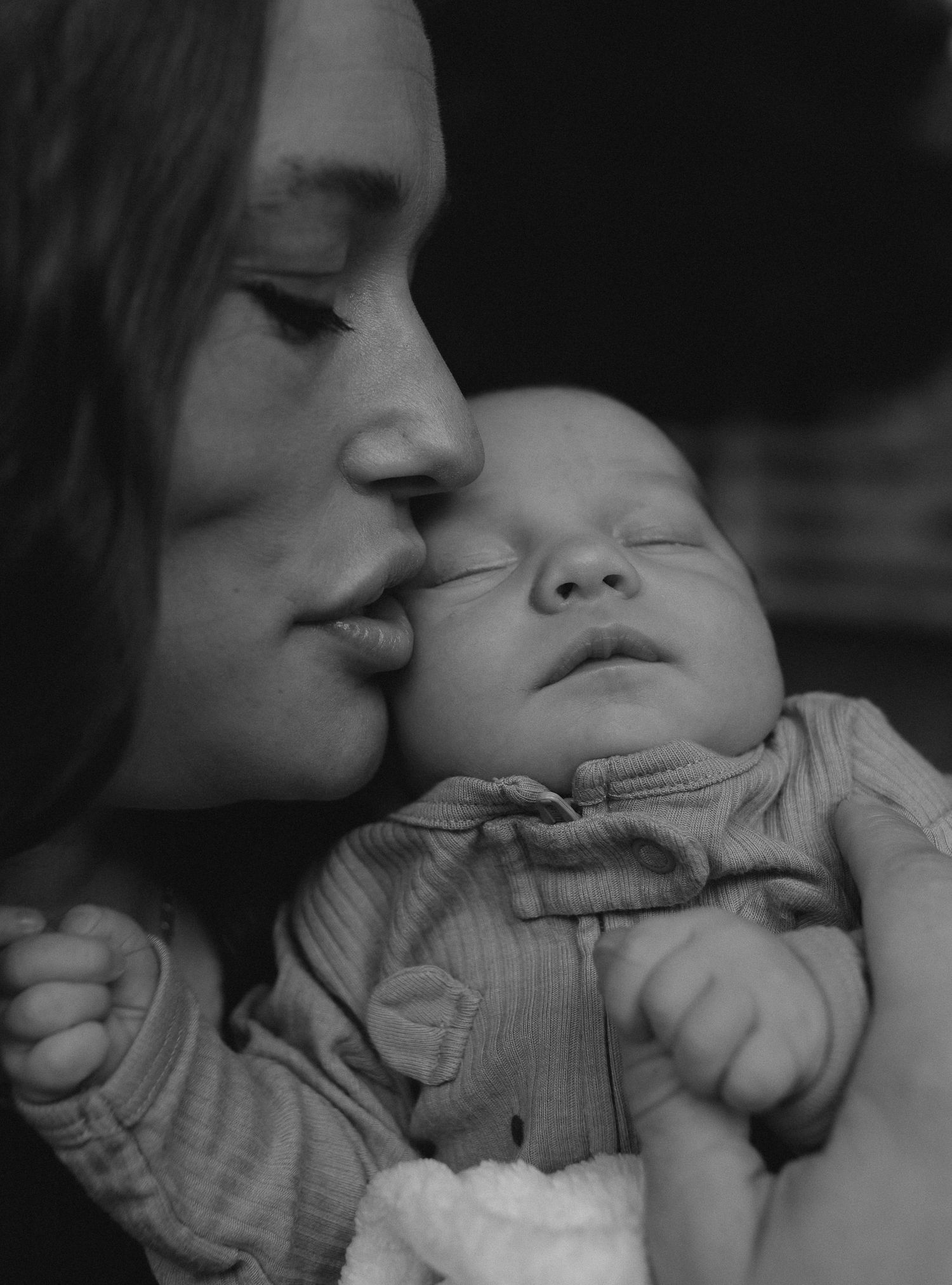 Black and white portrait of mother tenderly kissing her sleeping newborn baby held gently in her arms.