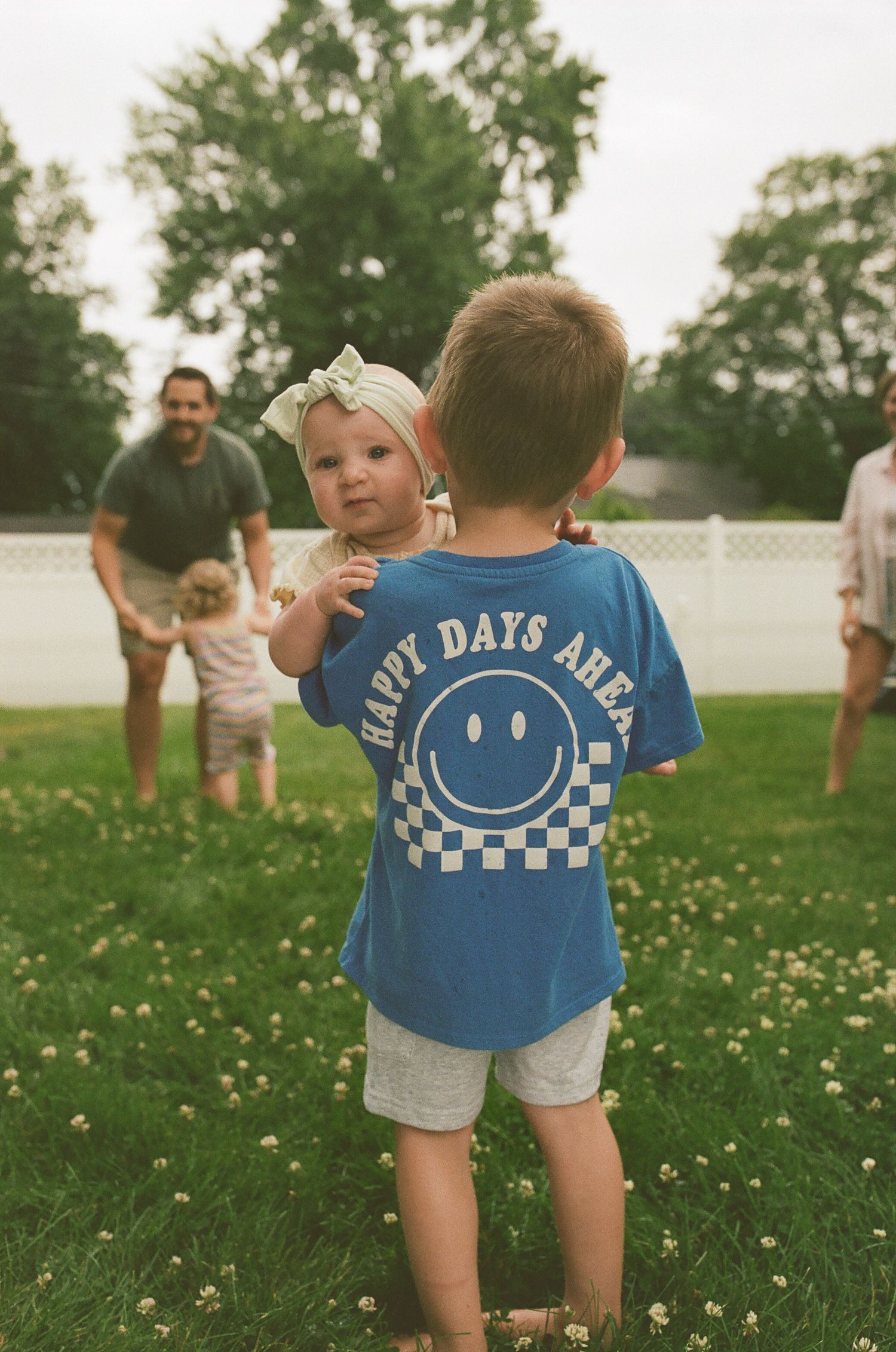 Boy in blue Happy Days Ahead shirt holding baby sister in sunny backyard.