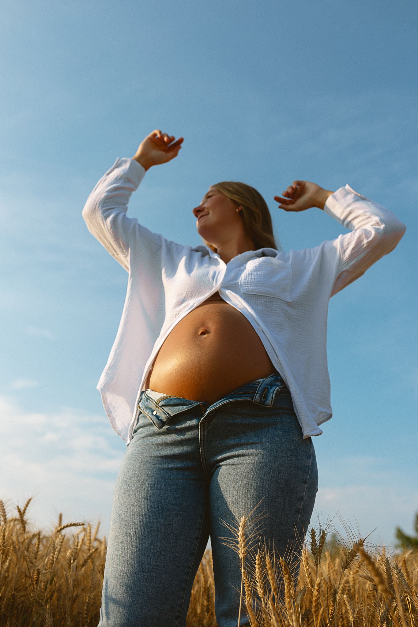 Pregnant woman in white shirt and jeans standing in golden wheat field under blue sky.