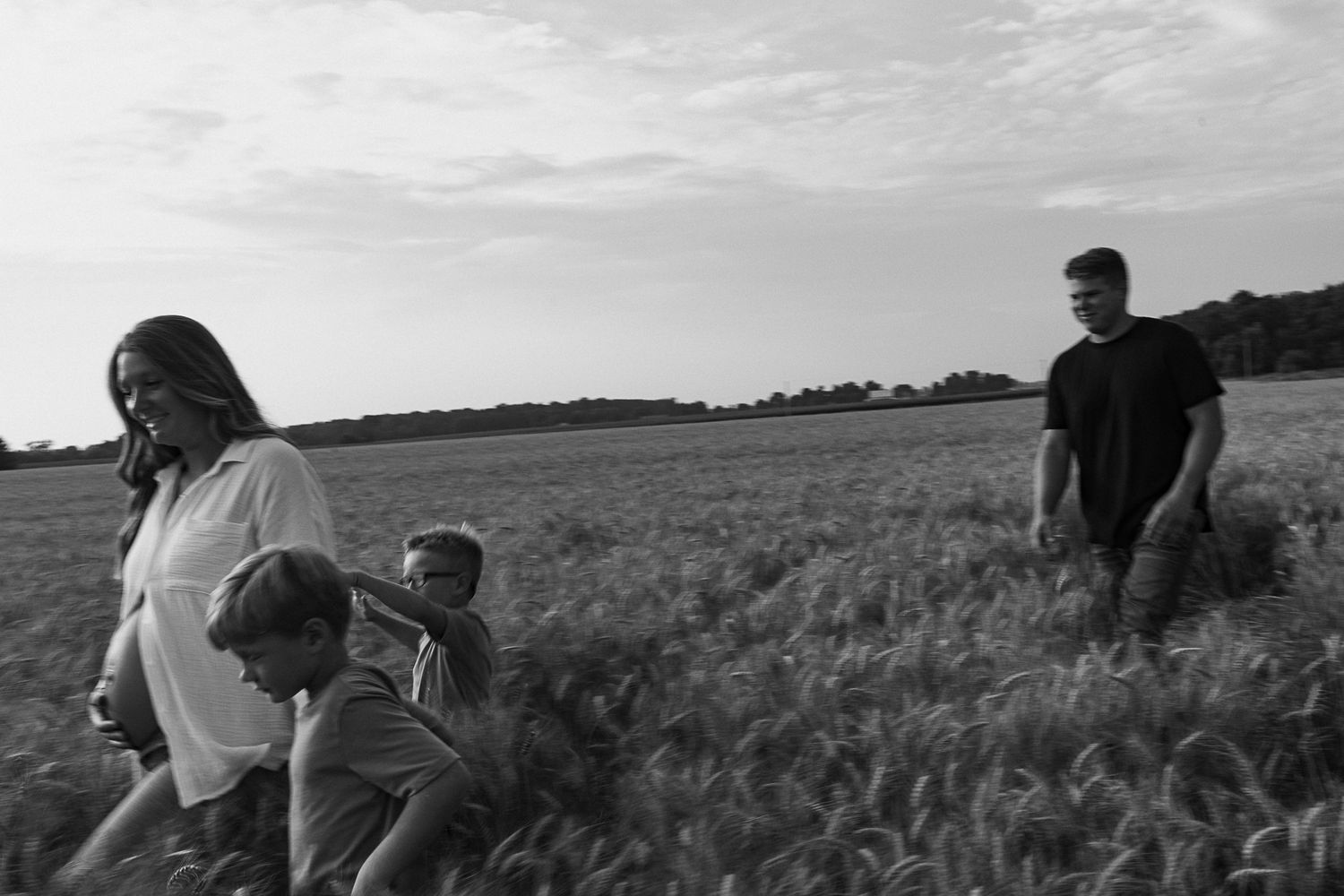 Black and white photo of family of four walking through open field under cloudy sky.