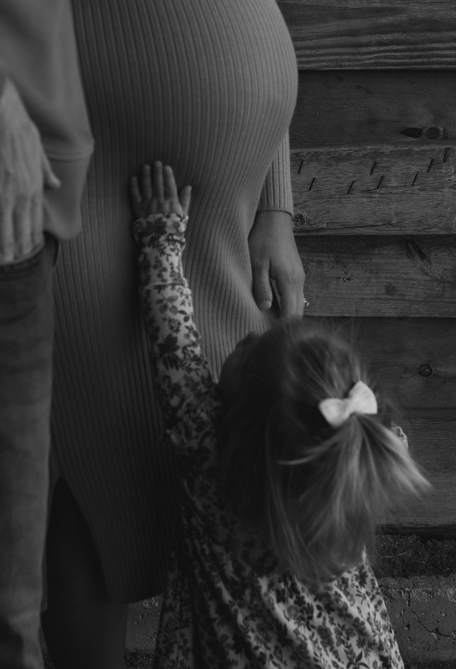Black and white image of toddler girl with bow touching mothers pregnant belly near barn.