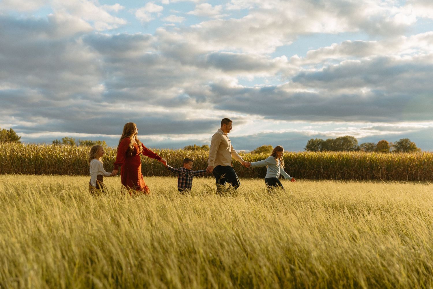 Family of five holding hands walking through a golden wheat field under a dramatic cloudy sky.