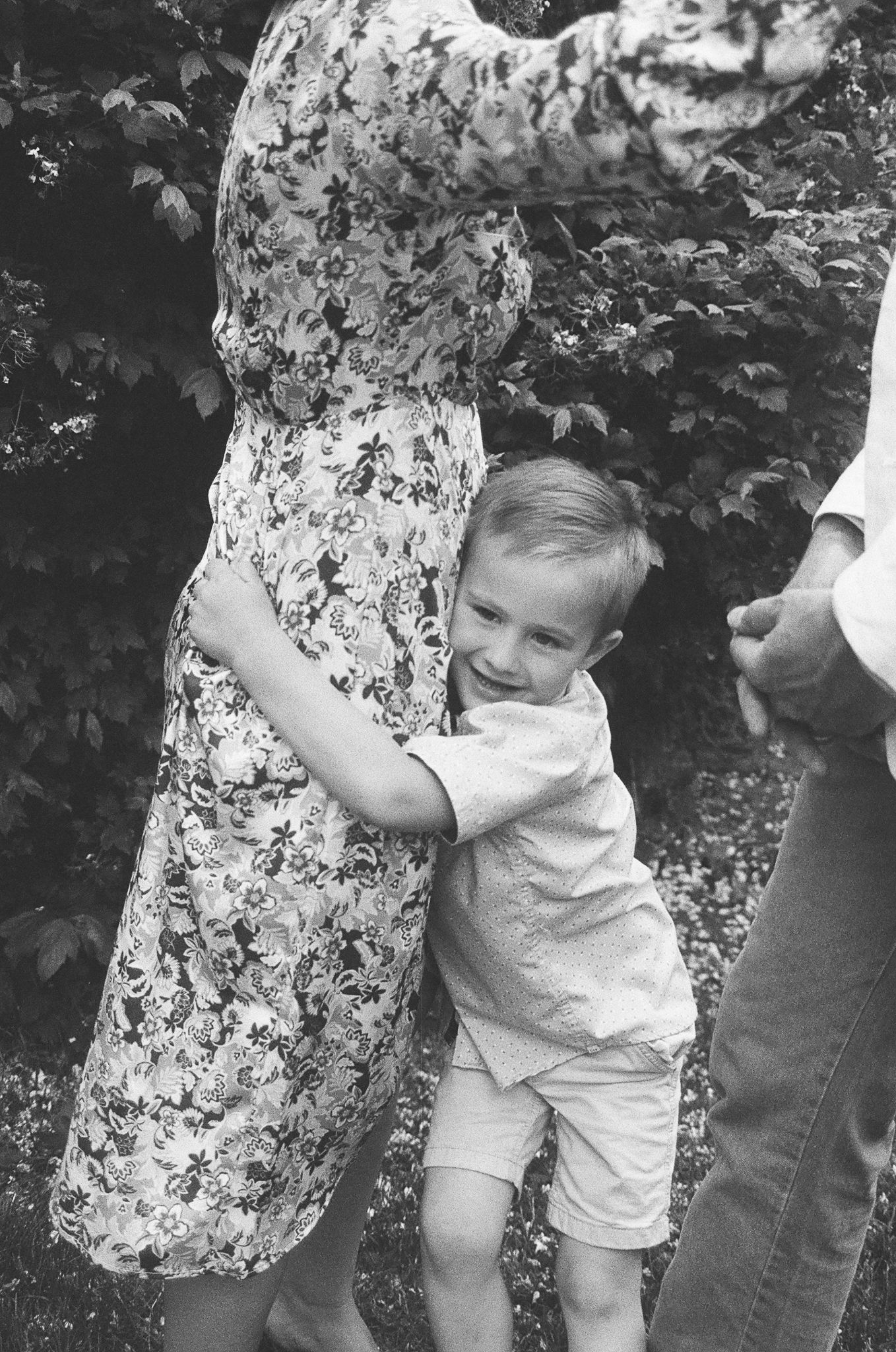 Black and white photo of a happy young boy hugging his mothers legs in a garden setting.