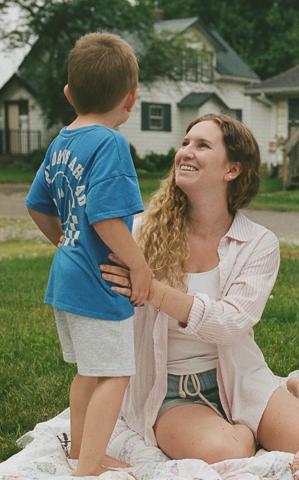 Smiling curly-haired mother sitting on grass interacting with her young son wearing a blue graphic t-shirt outdoors.