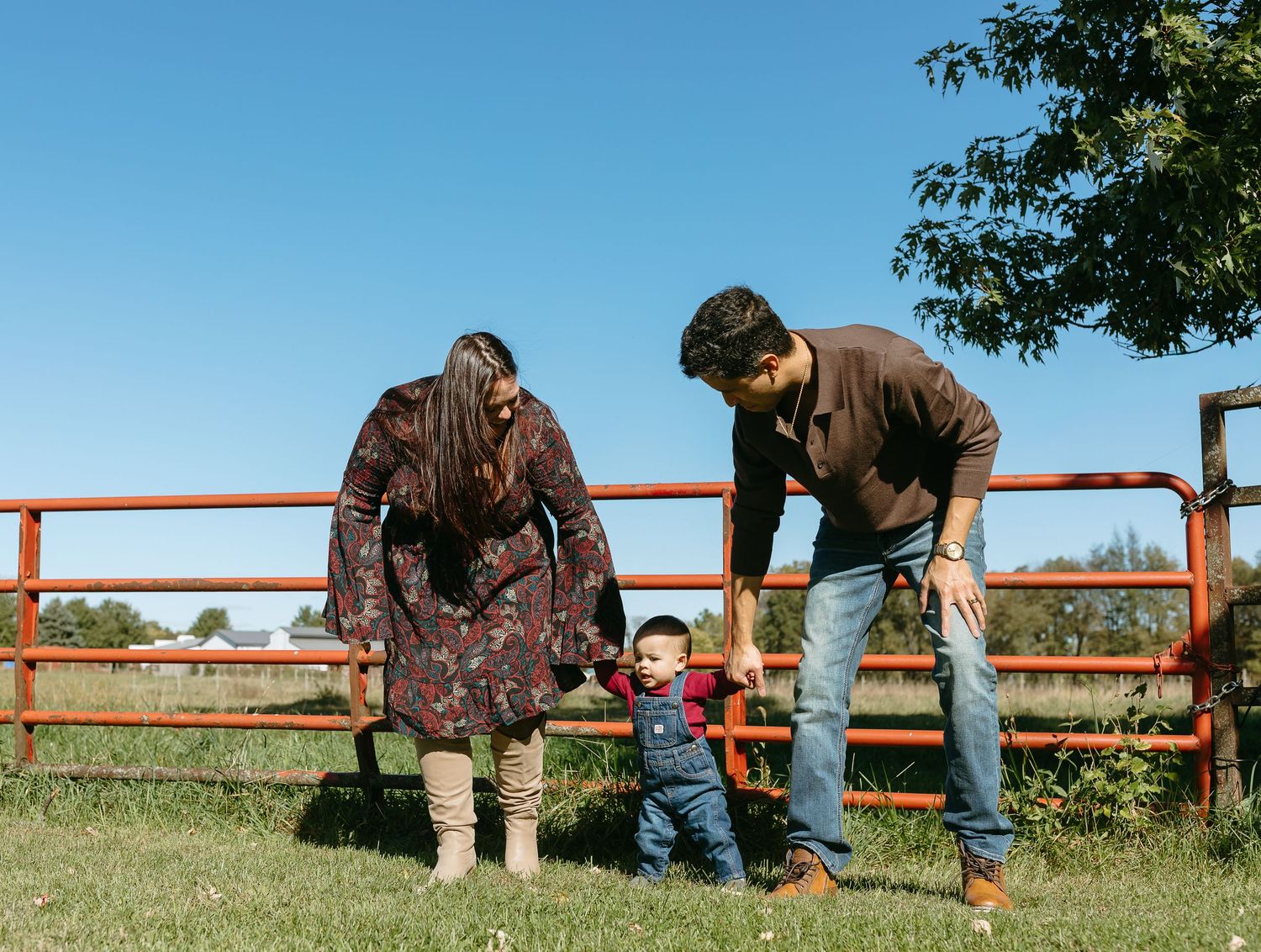 Young couple encouraging their toddler to walk by a red farm fence under a clear blue sky.