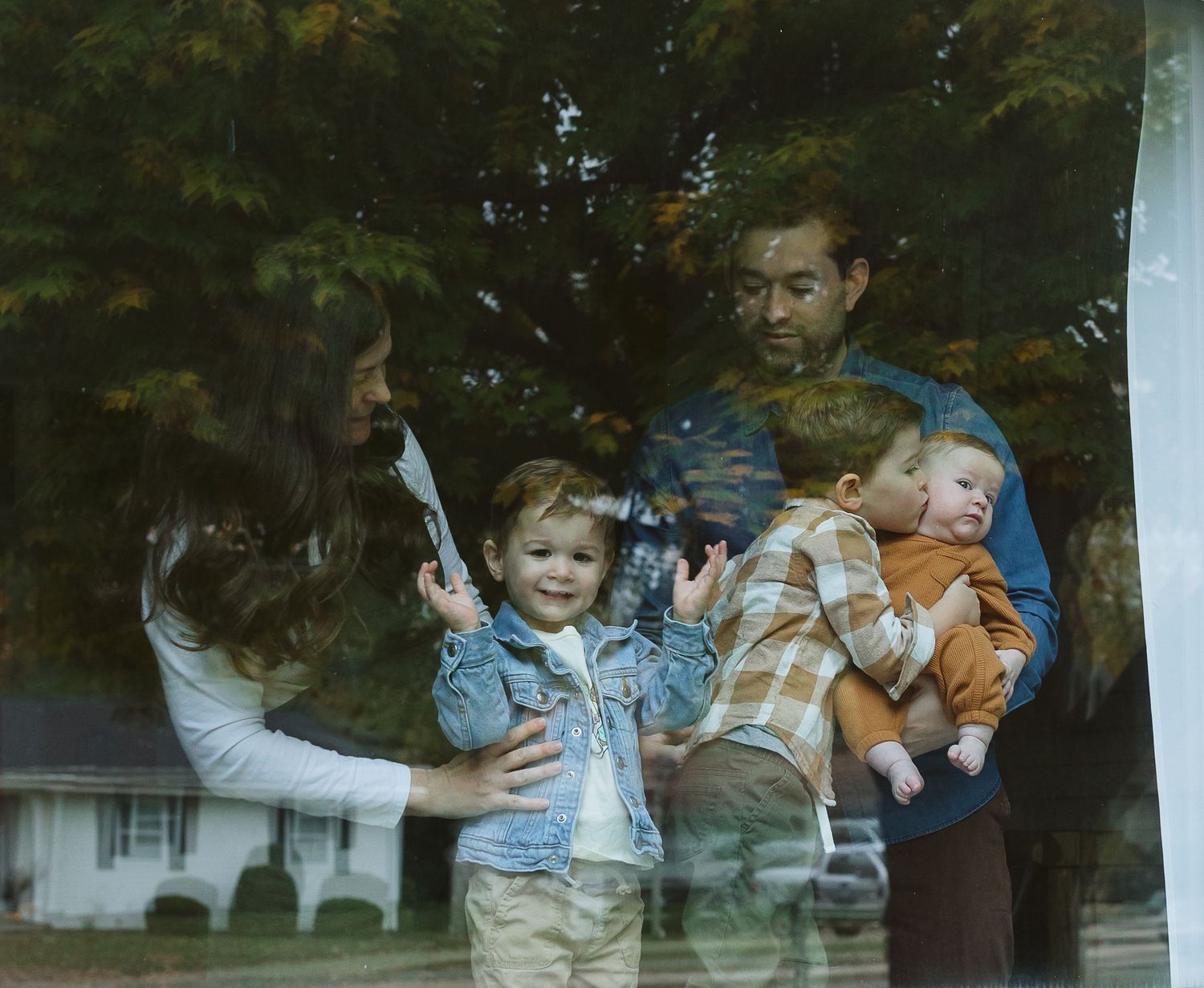 Family of four seen through a window, parents holding two young children outdoors.