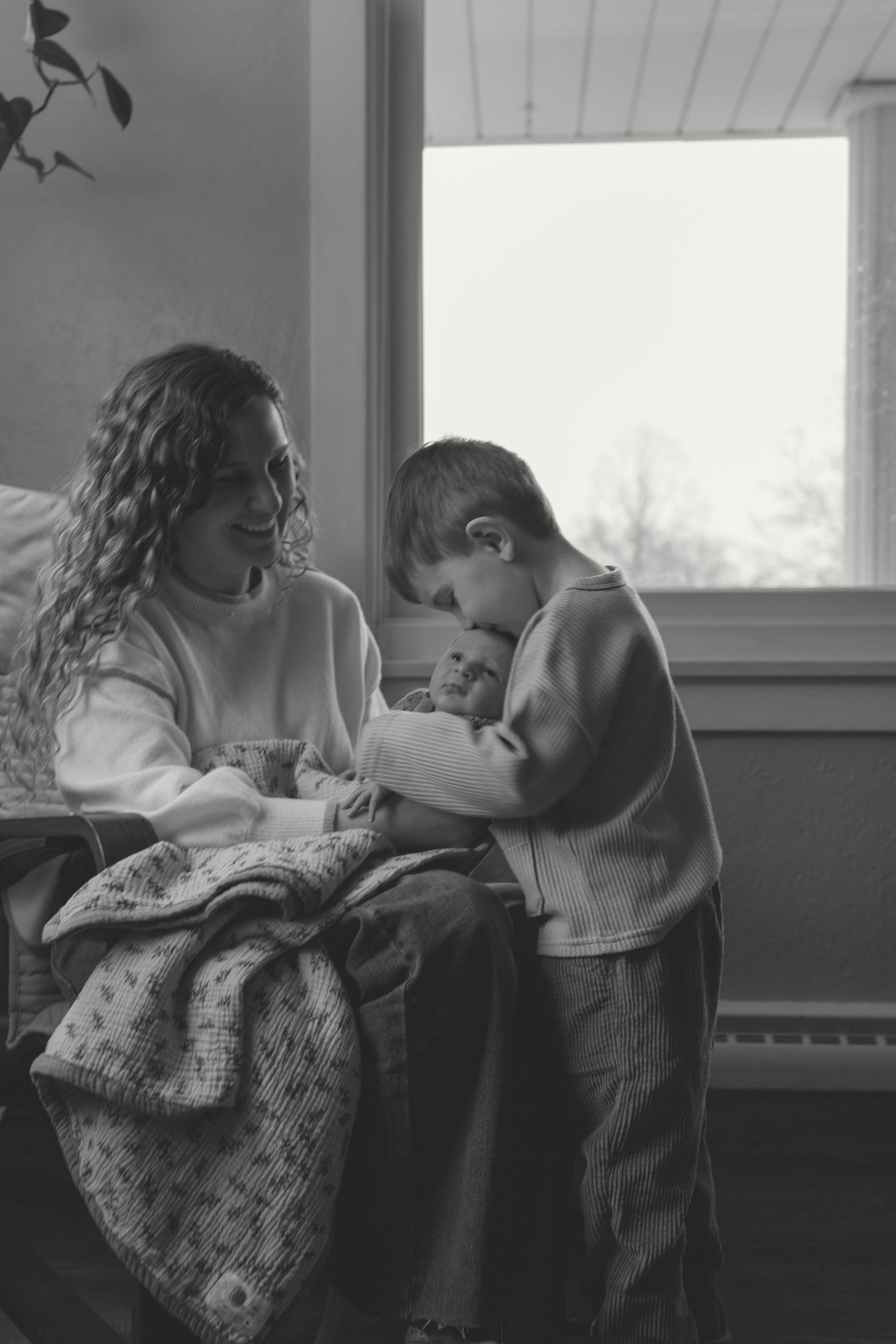 Black and white photo of a mother smiling as young boy holds newborn baby indoors.