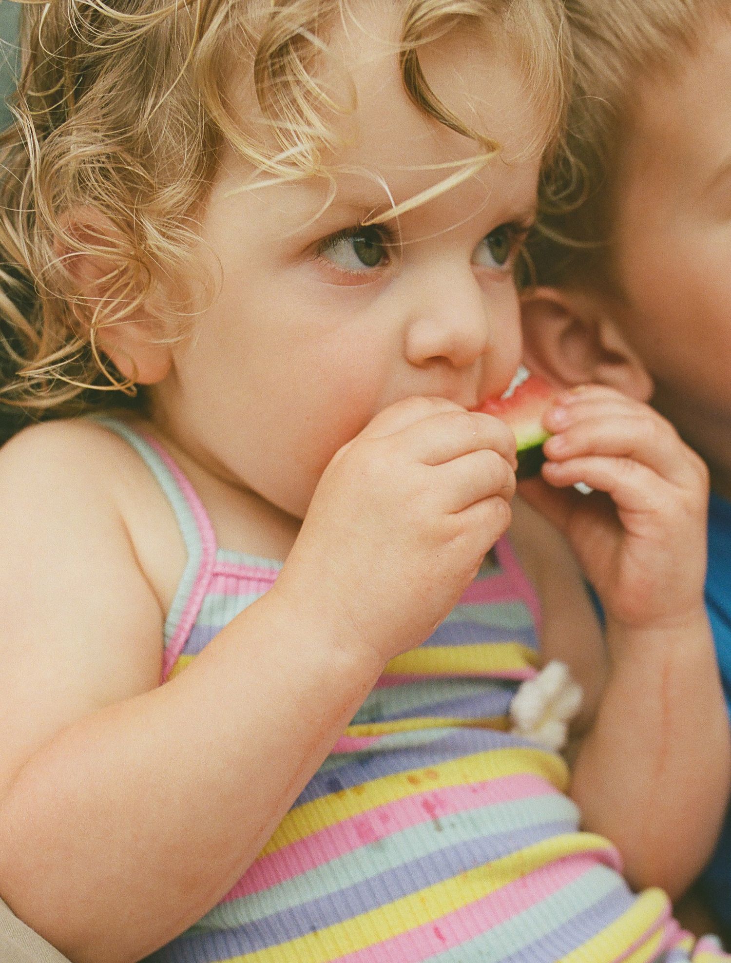 Adorable curly-haired toddler girl in colorful striped top eating a piece of fresh watermelon outdoors.