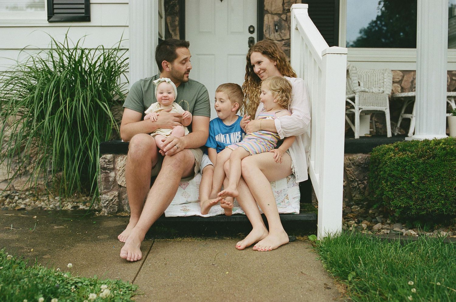Happy family of five sitting together on front porch steps of suburban home.