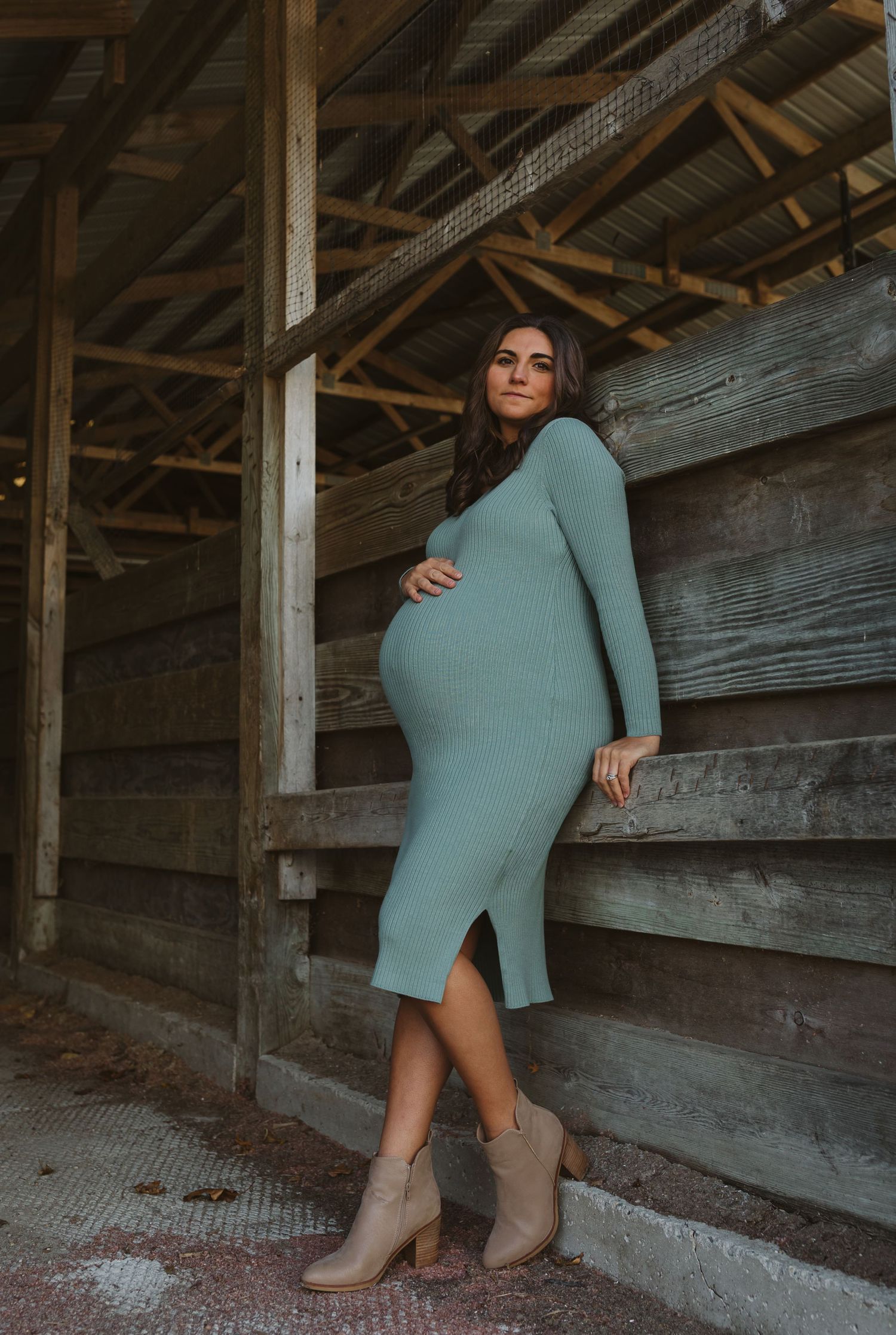 Pregnant brunette woman in fitted sage green dress poses against rustic wooden barn wall.