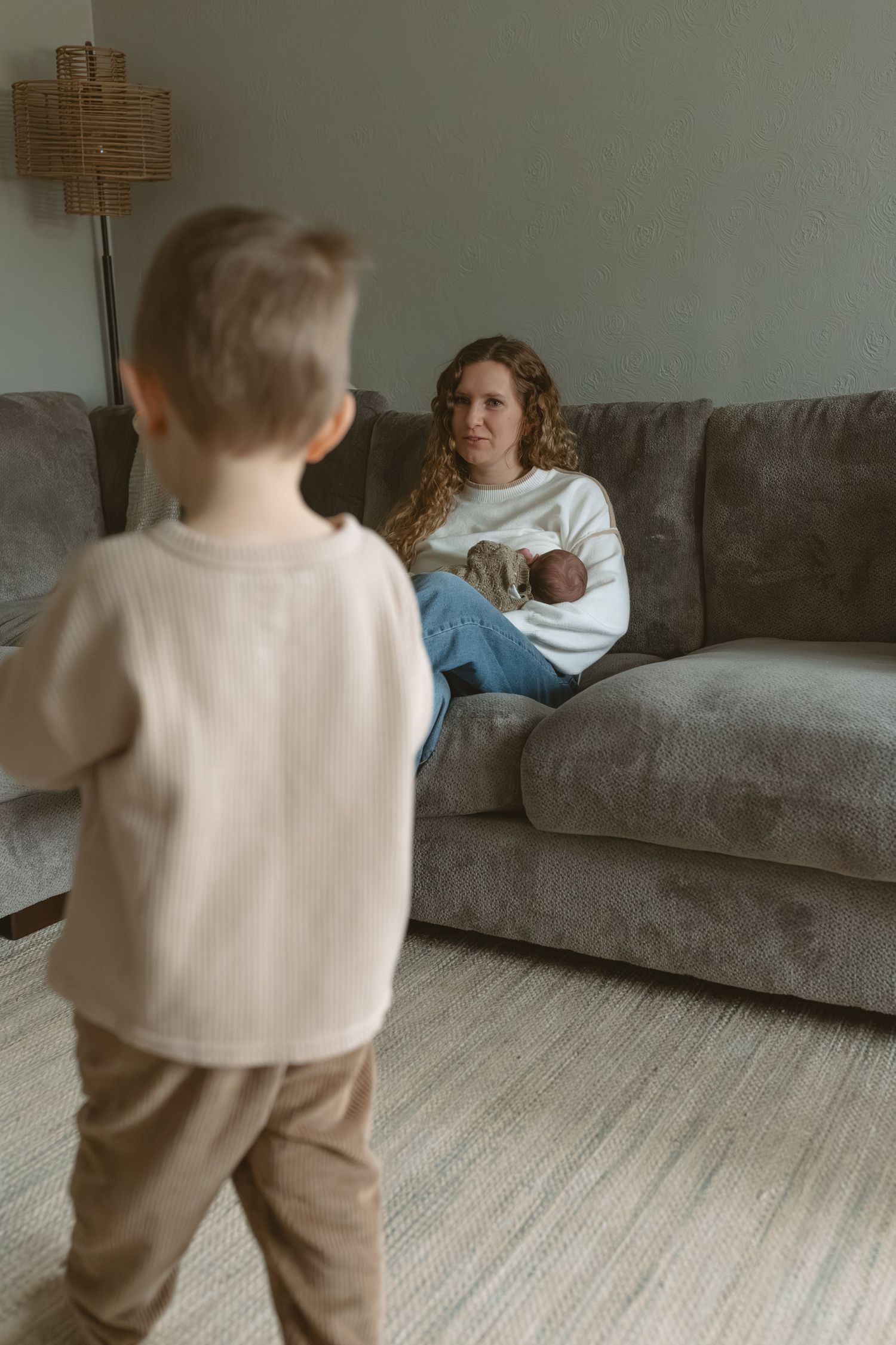 Curly-haired mother holds newborn baby on gray sofa while young toddler son stands in foreground of cozy living room.