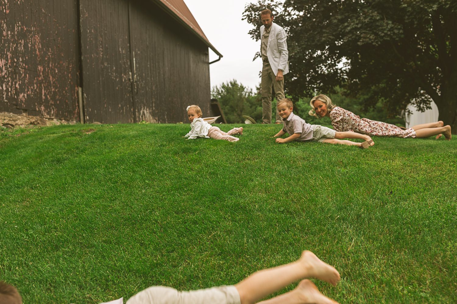 Children playing and rolling down a grassy hill beside a rustic barn on a cloudy day.