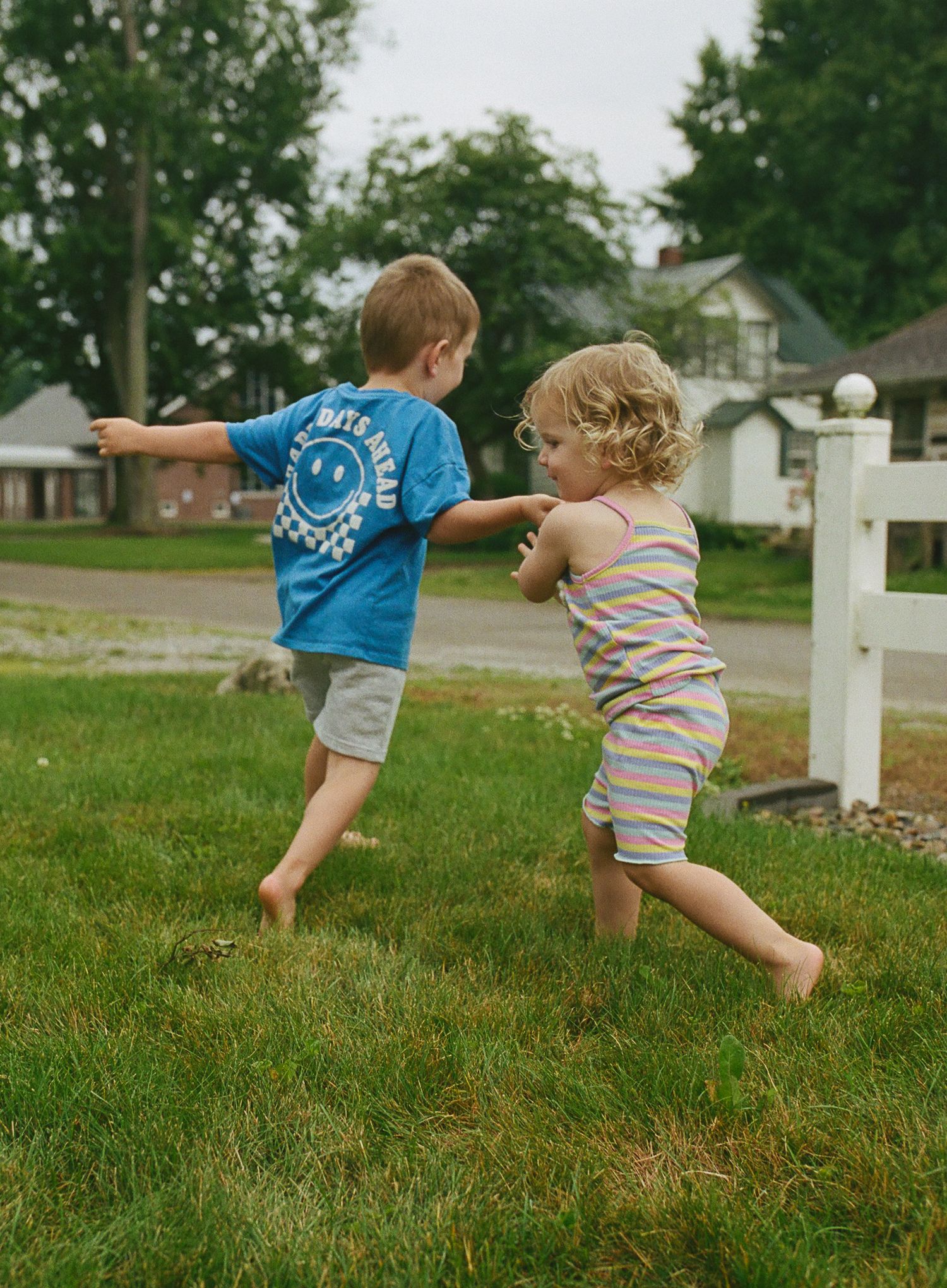 Two barefoot children playing and running together on green grass in a suburban backyard on a summer day.