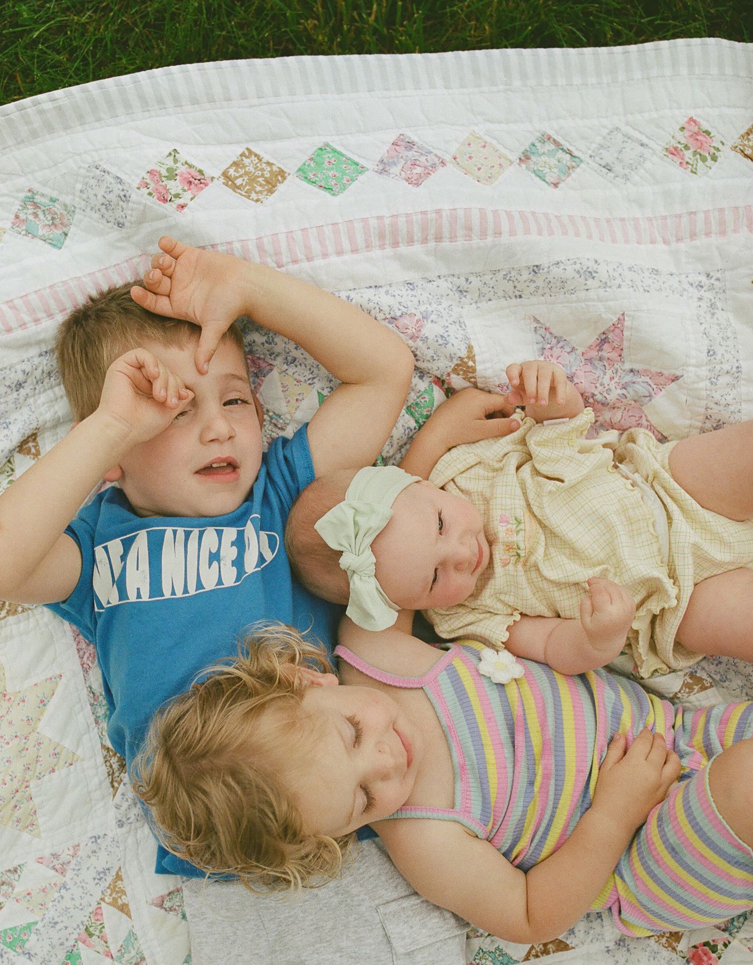 Three young siblings lying together on colorful patchwork quilt outdoors.
