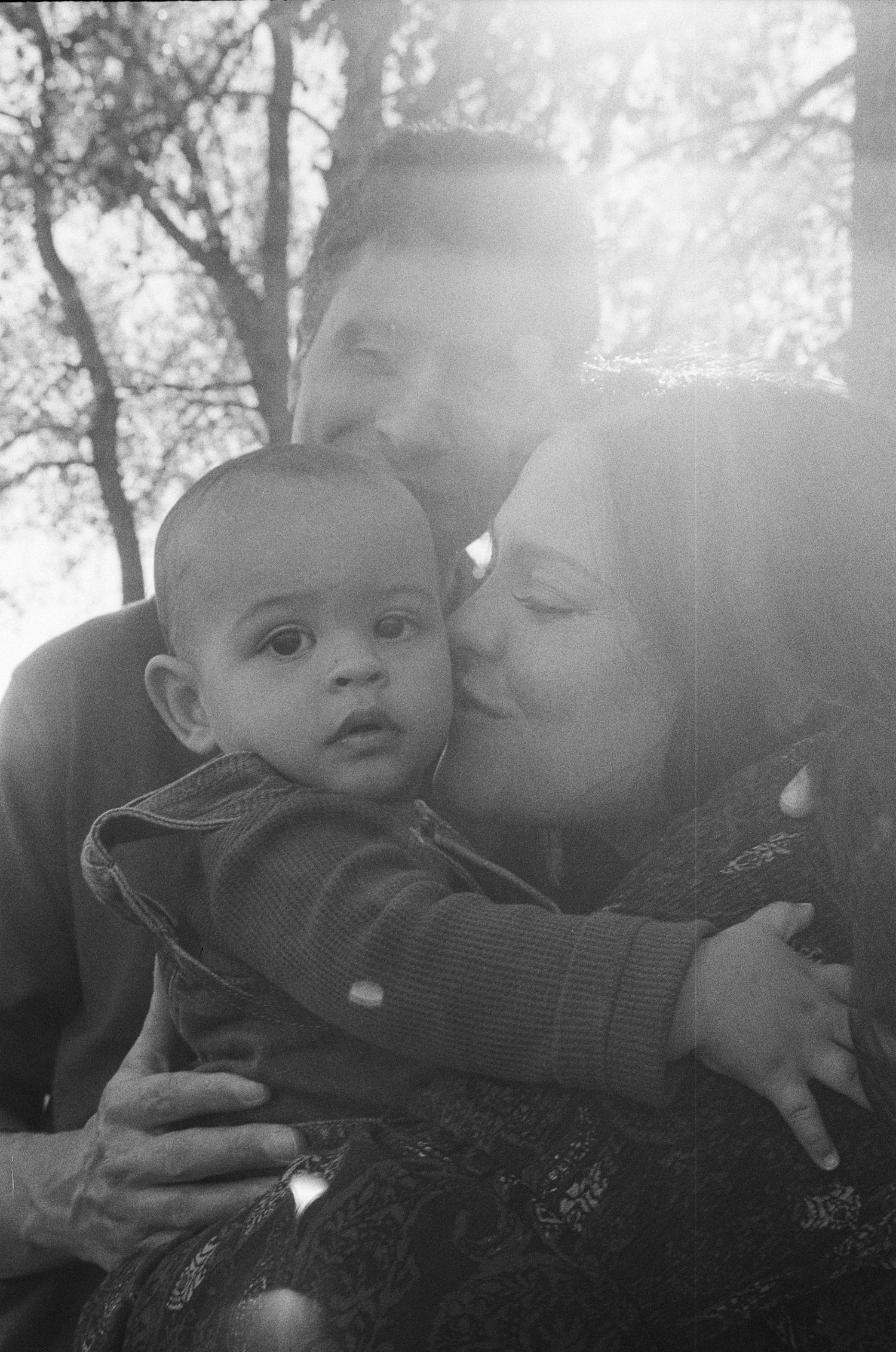 Black and white family portrait of parents kissing their infant baby outdoors.