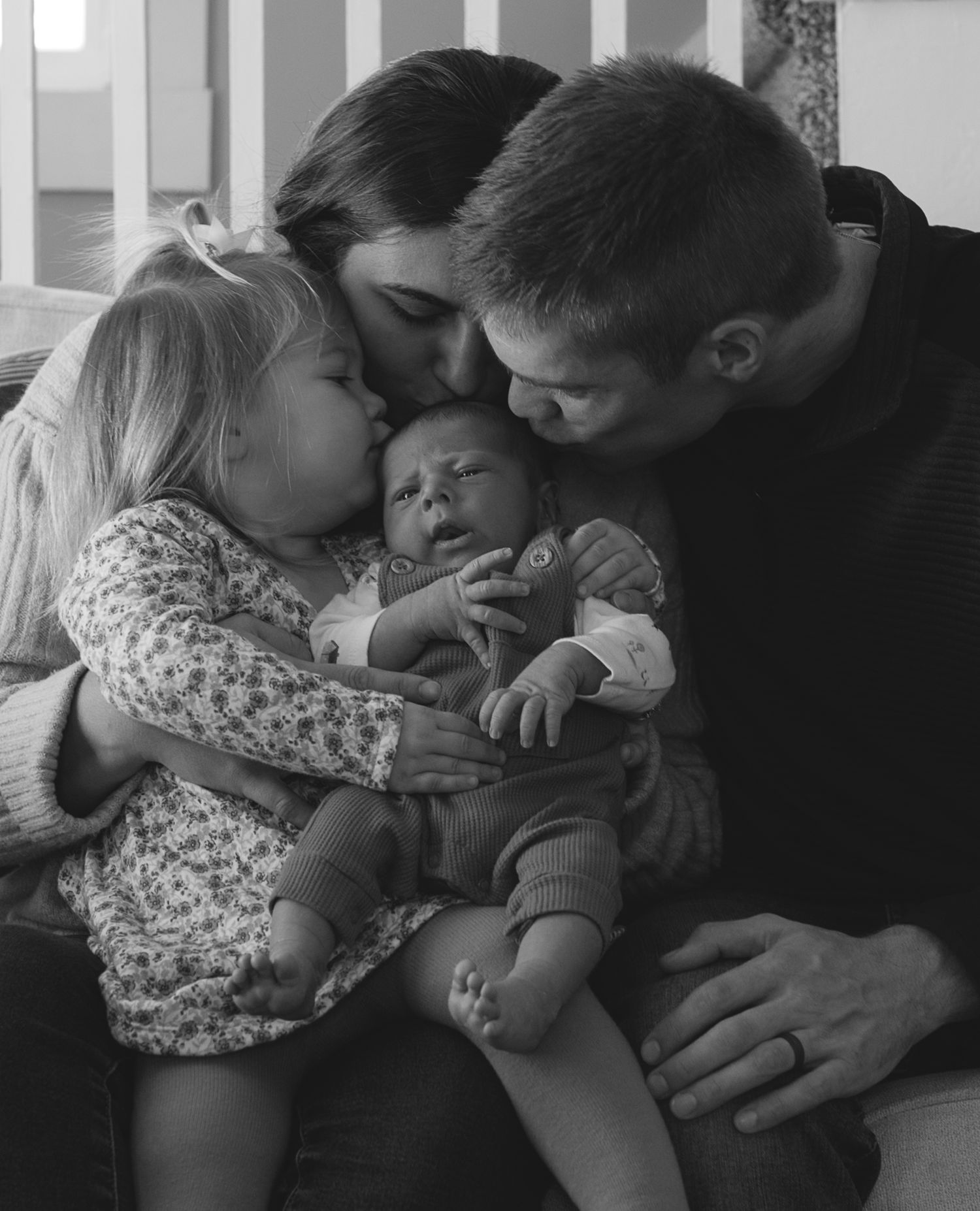 Parents and toddler kissing newborn baby in tender black and white family portrait.
