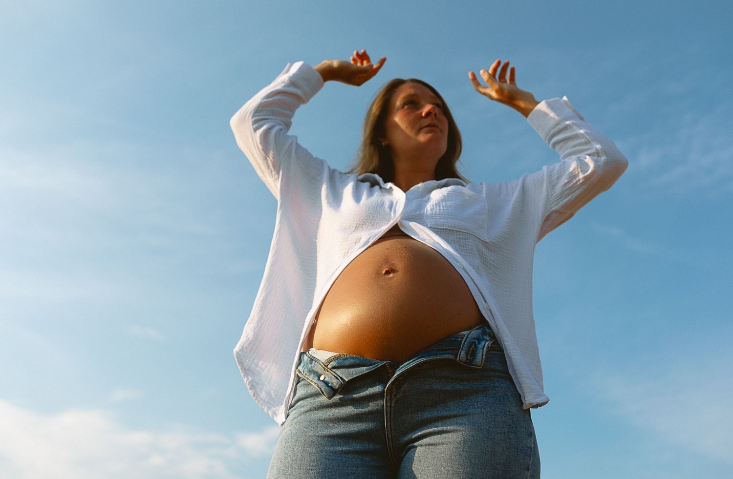 Pregnant woman in white shirt and jeans posing outdoors against blue sky, showing baby bump.