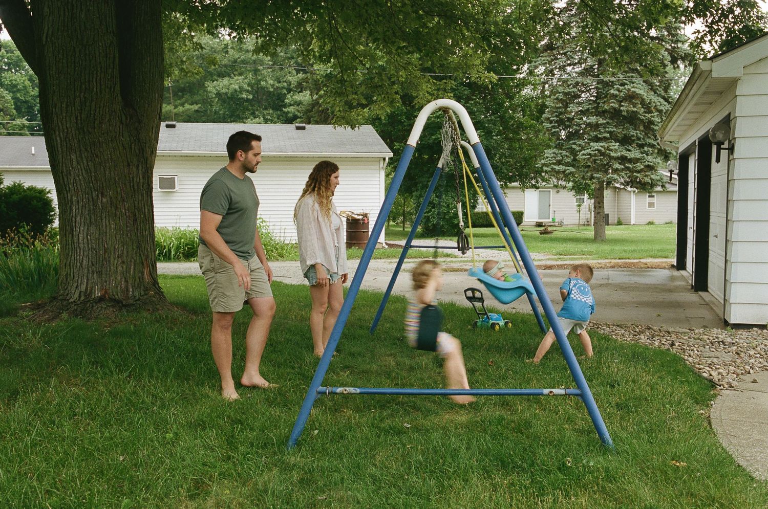 Parents watching children play on backyard swing set on sunny summer day.