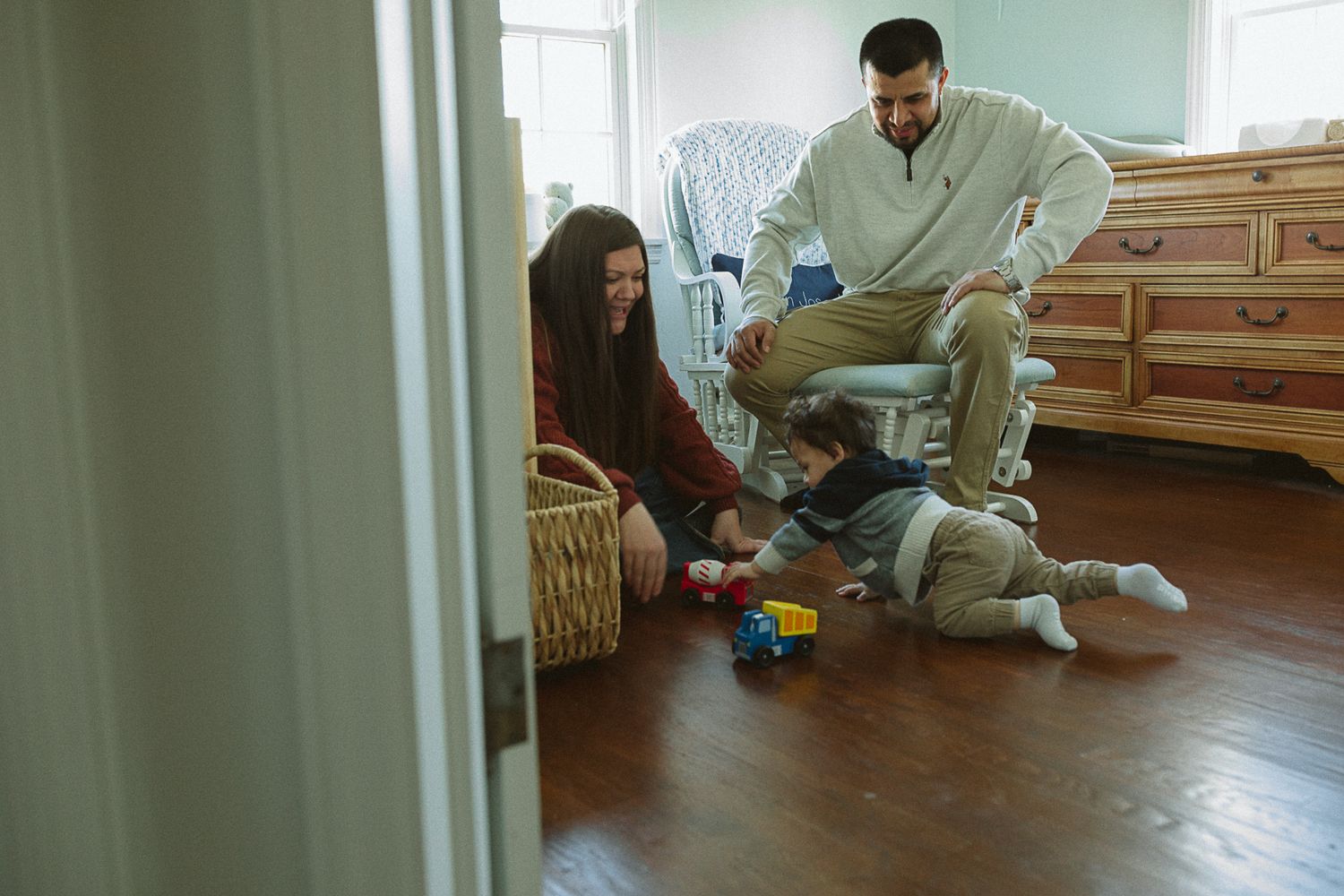 Young family plays with baby on hardwood floor in cozy nursery room.