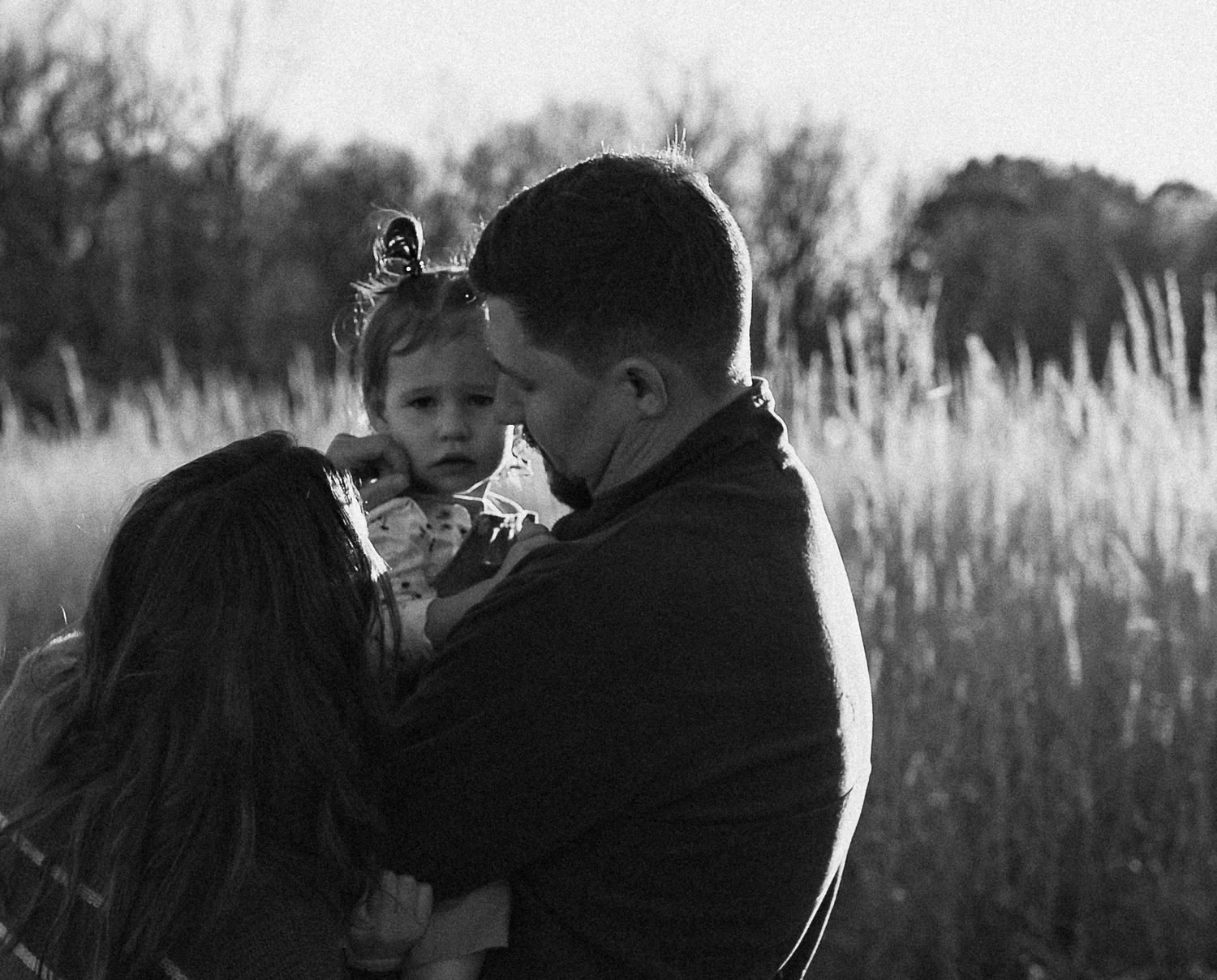 Black and white photo of parents lovingly kissing their young daughter in a grassy field.