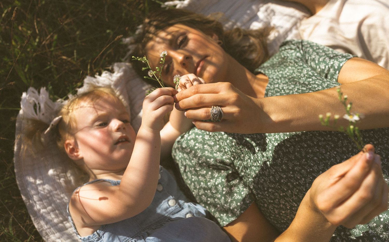Mother and blonde toddler lay on blanket in sunny grass examining a small flower together.