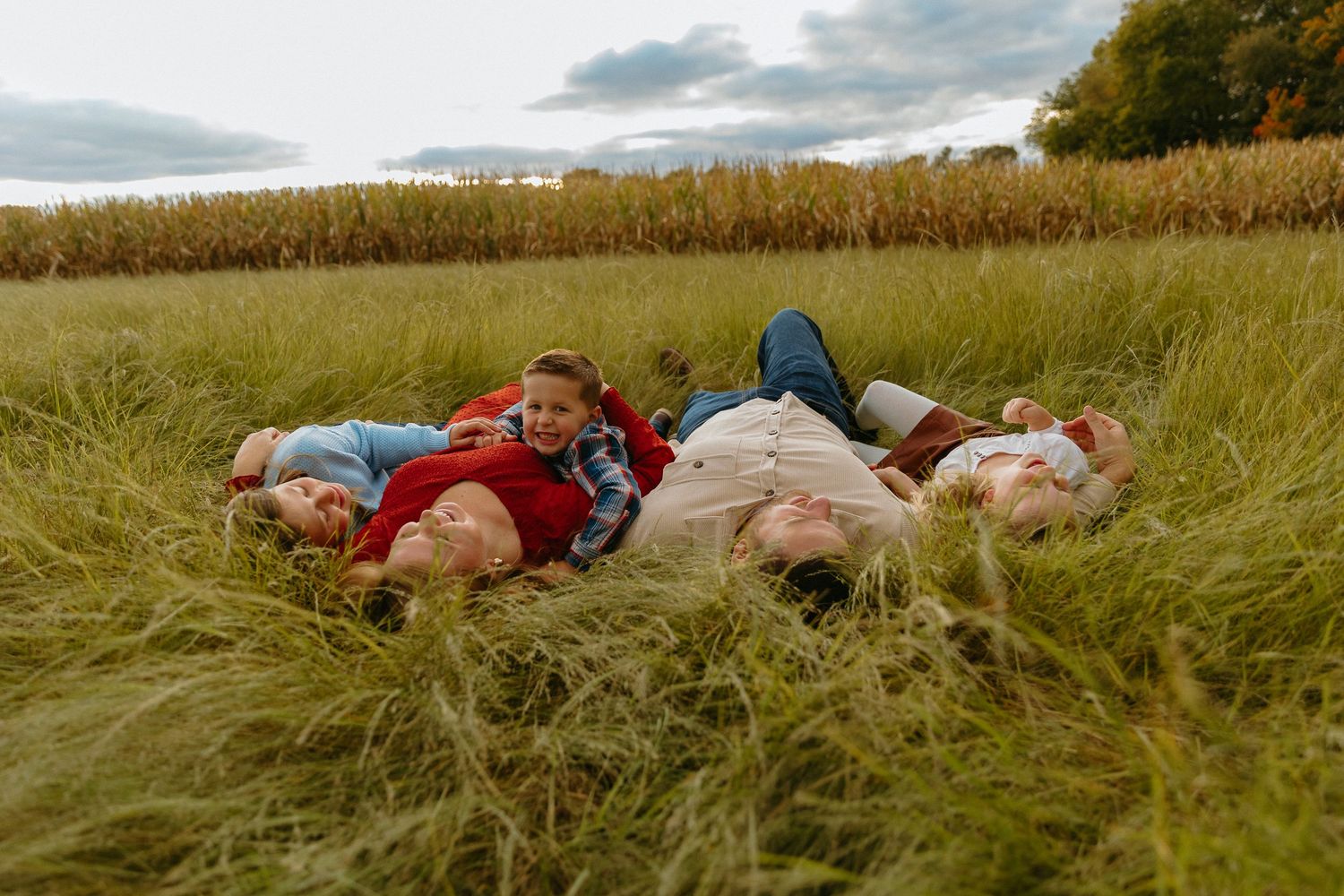 Family of four lying in tall grass field near cornfield on cloudy day.