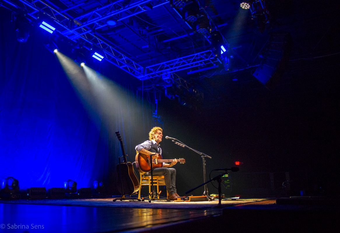 Solo musician performing acoustic guitar set on stage bathed in dramatic blue stage lighting.