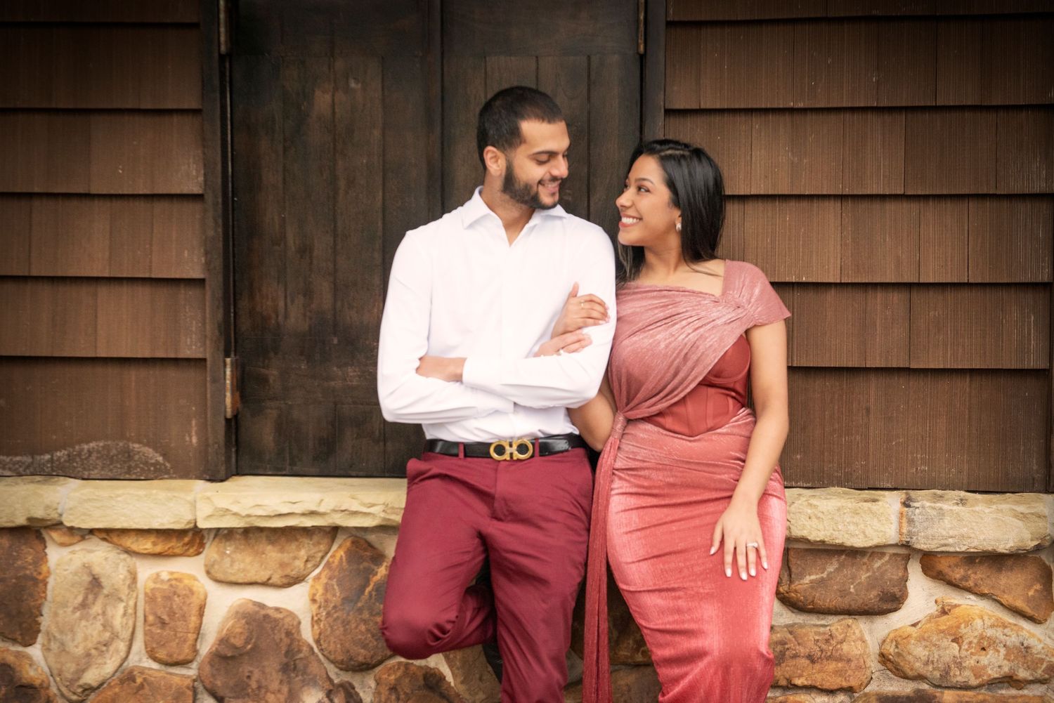 Couple in matching pink and burgundy outfits smiling at each other by a wooden building.