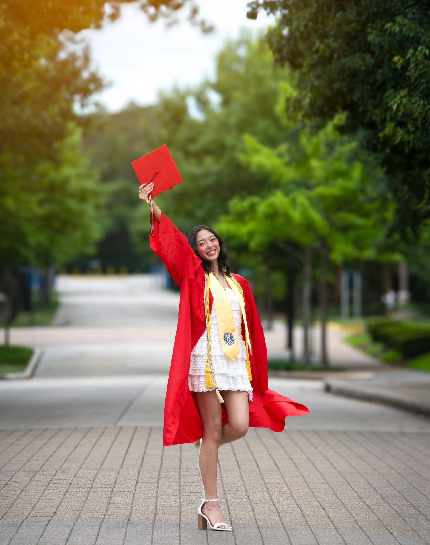 Young woman in red graduation gown and cap celebrating outdoors on a tree-lined campus walkway.