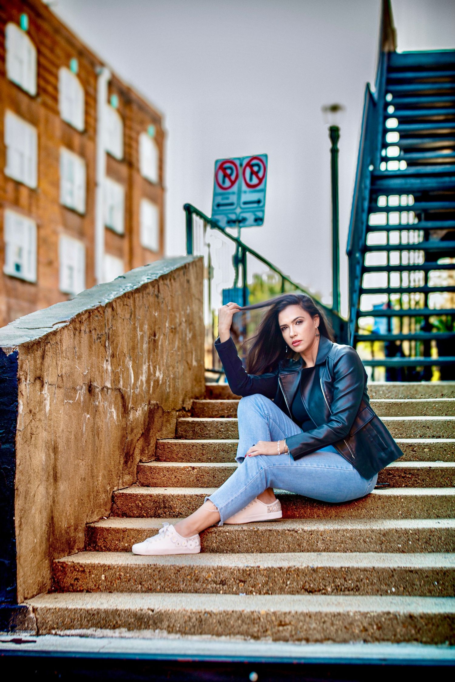 Trendy woman in black leather jacket and jeans sitting on outdoor stairs with urban brick building backdrop.