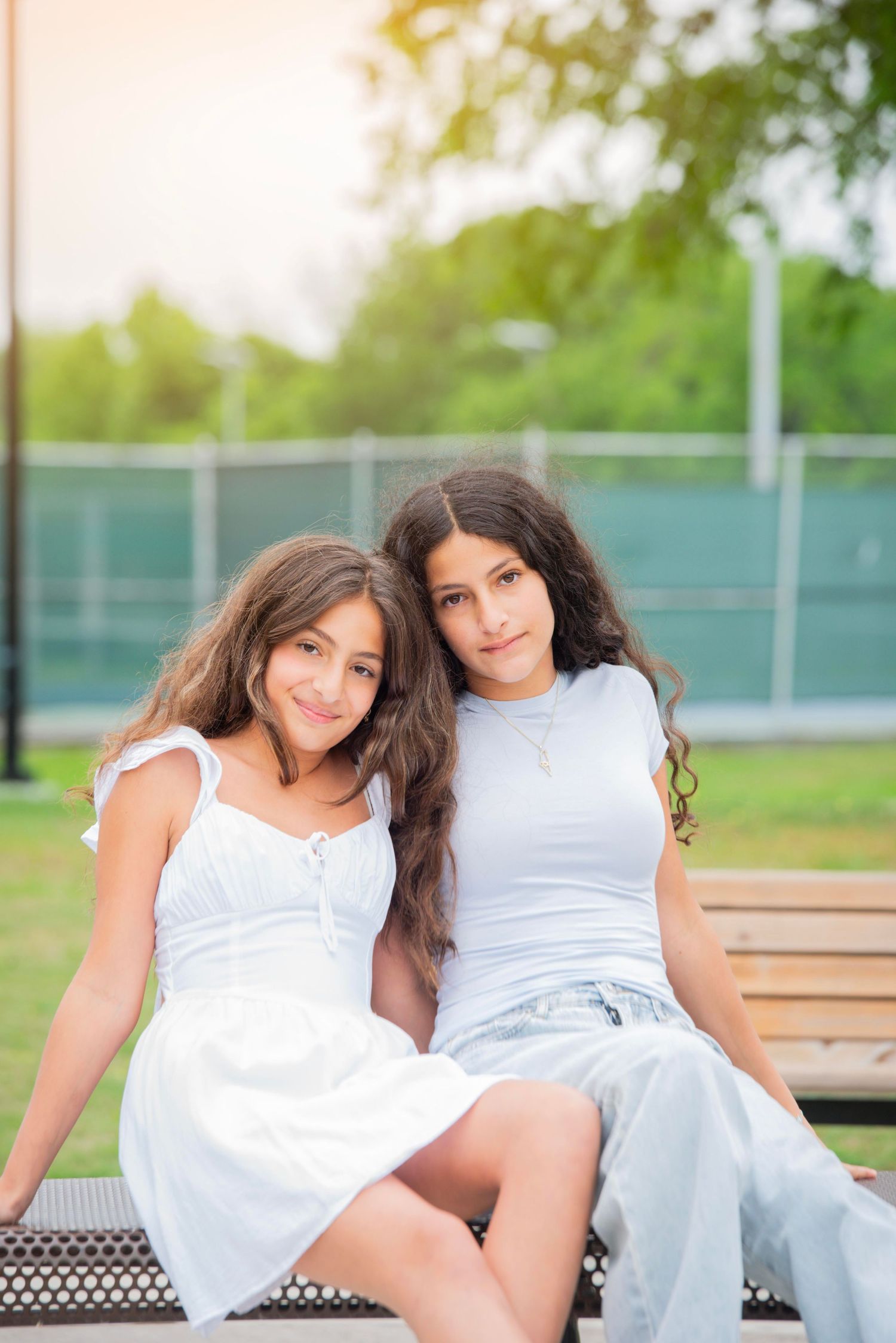 Two young girls with long dark hair sit together on a bench in a park, wearing white outfits, smiling at the camera.