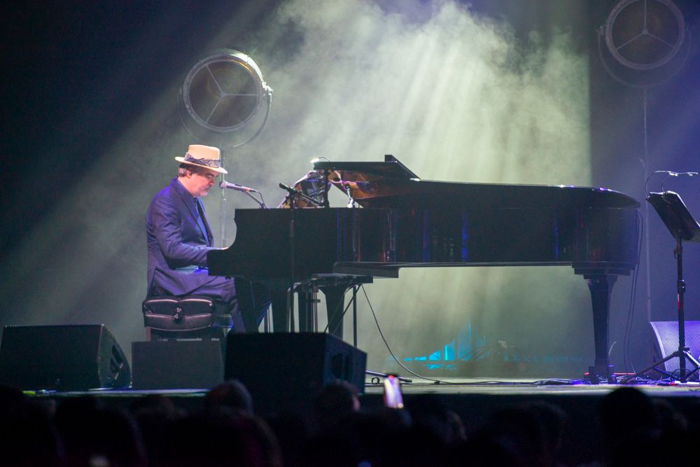 Musician in purple hat playing grand piano on a smoky, atmospheric concert stage.