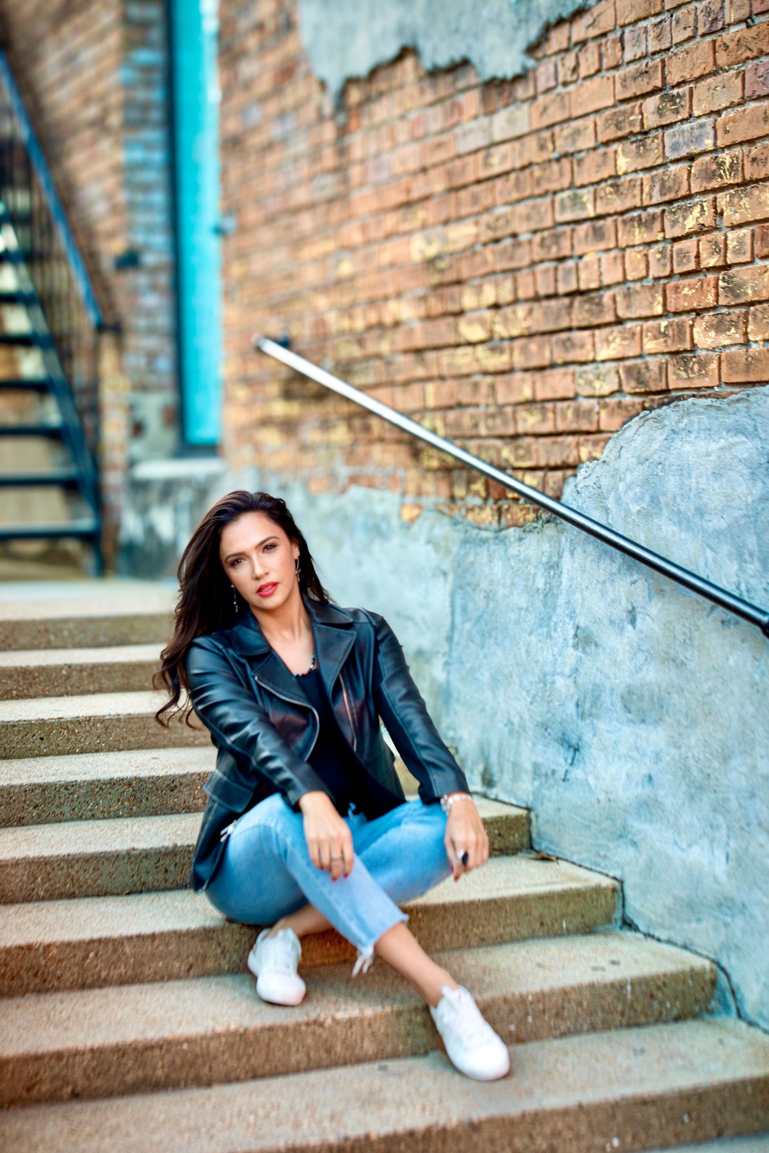 Young woman in black leather jacket and jeans sitting on urban brick staircase with metal railing.