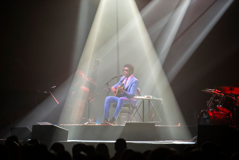 Musician performs solo on stage under dramatic spotlight, playing acoustic guitar while seated, surrounded by concert lighting.