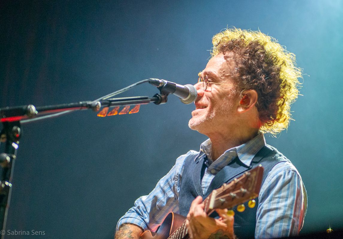 Curly-haired male guitarist smiling while performing at a live concert with microphone.