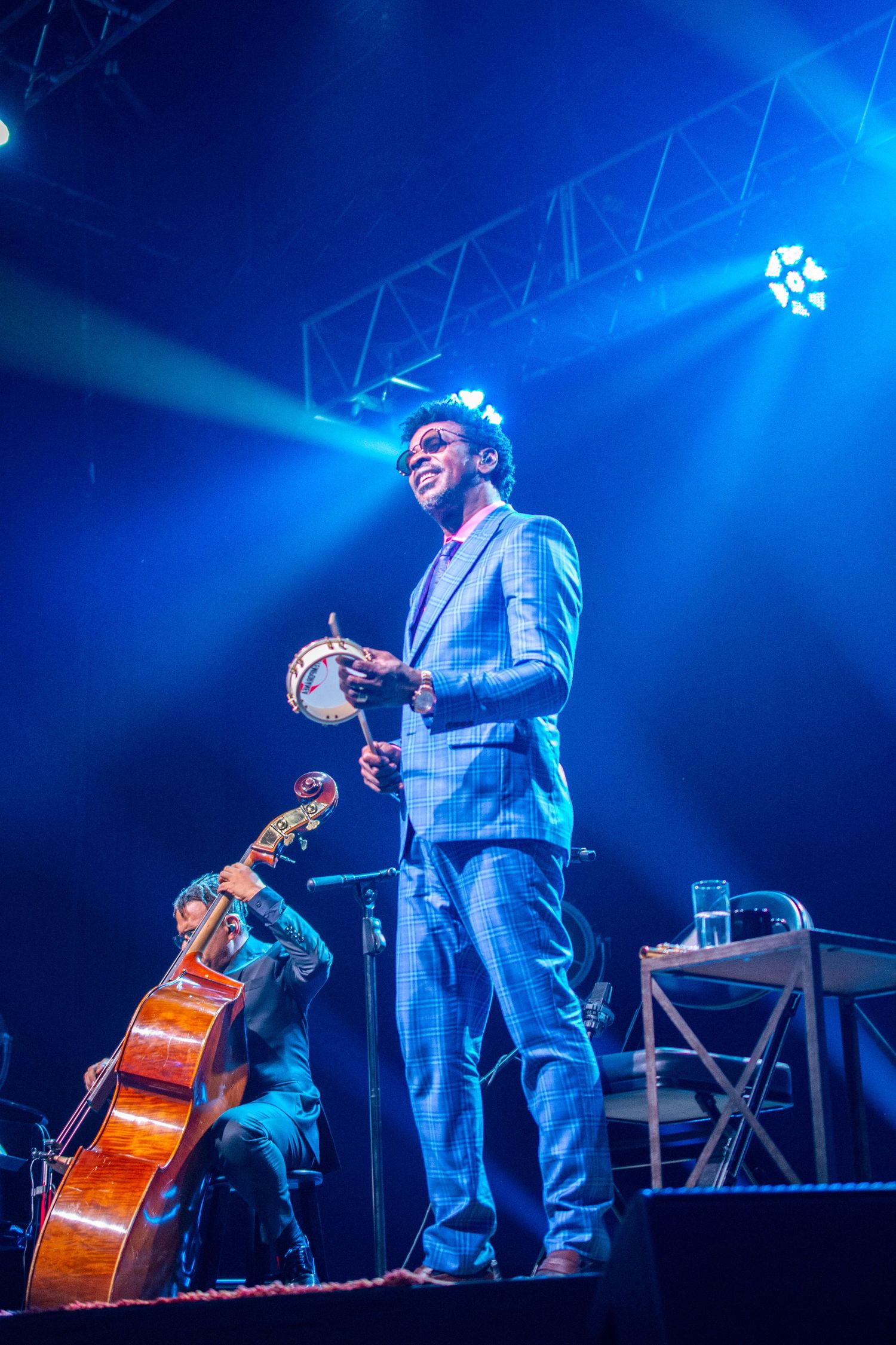 Male performer in blue suit playing tambourine on stage with dramatic blue lighting.
