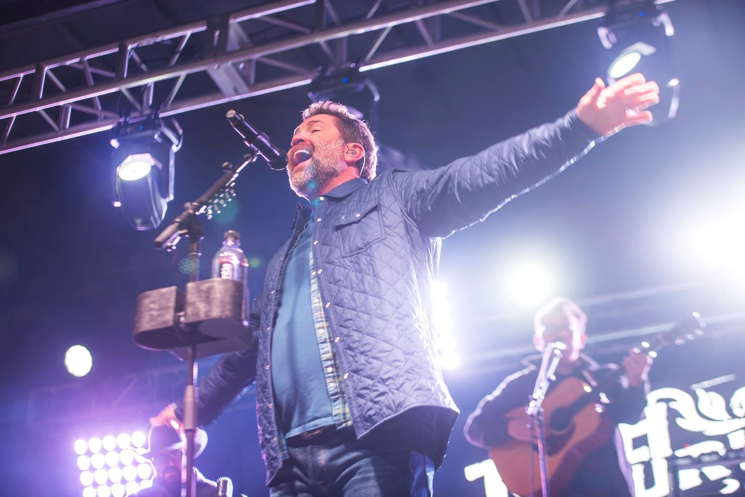 Energetic male vocalist performing on concert stage with arms raised under bright stage lights.