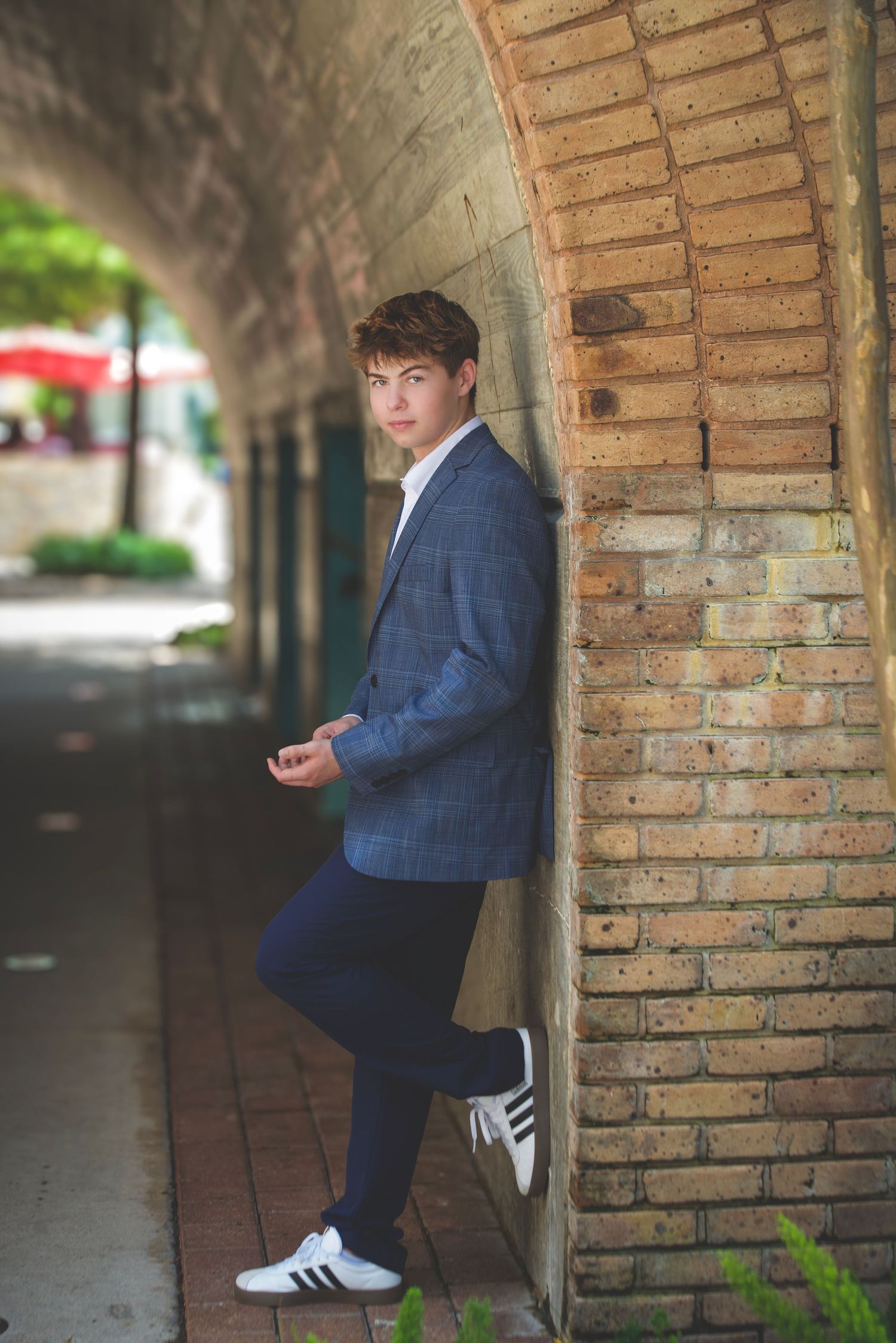 Young man in blue blazer and Adidas sneakers leaning against brick wall in an urban tunnel archway.