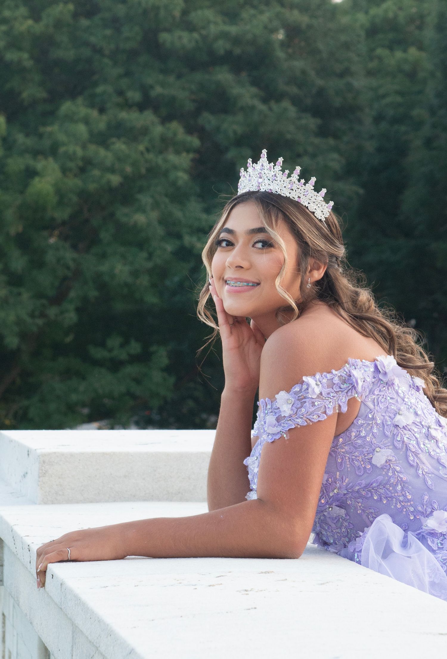 Smiling young woman wearing a crystal tiara and lavender floral gown leaning on a white balustrade.