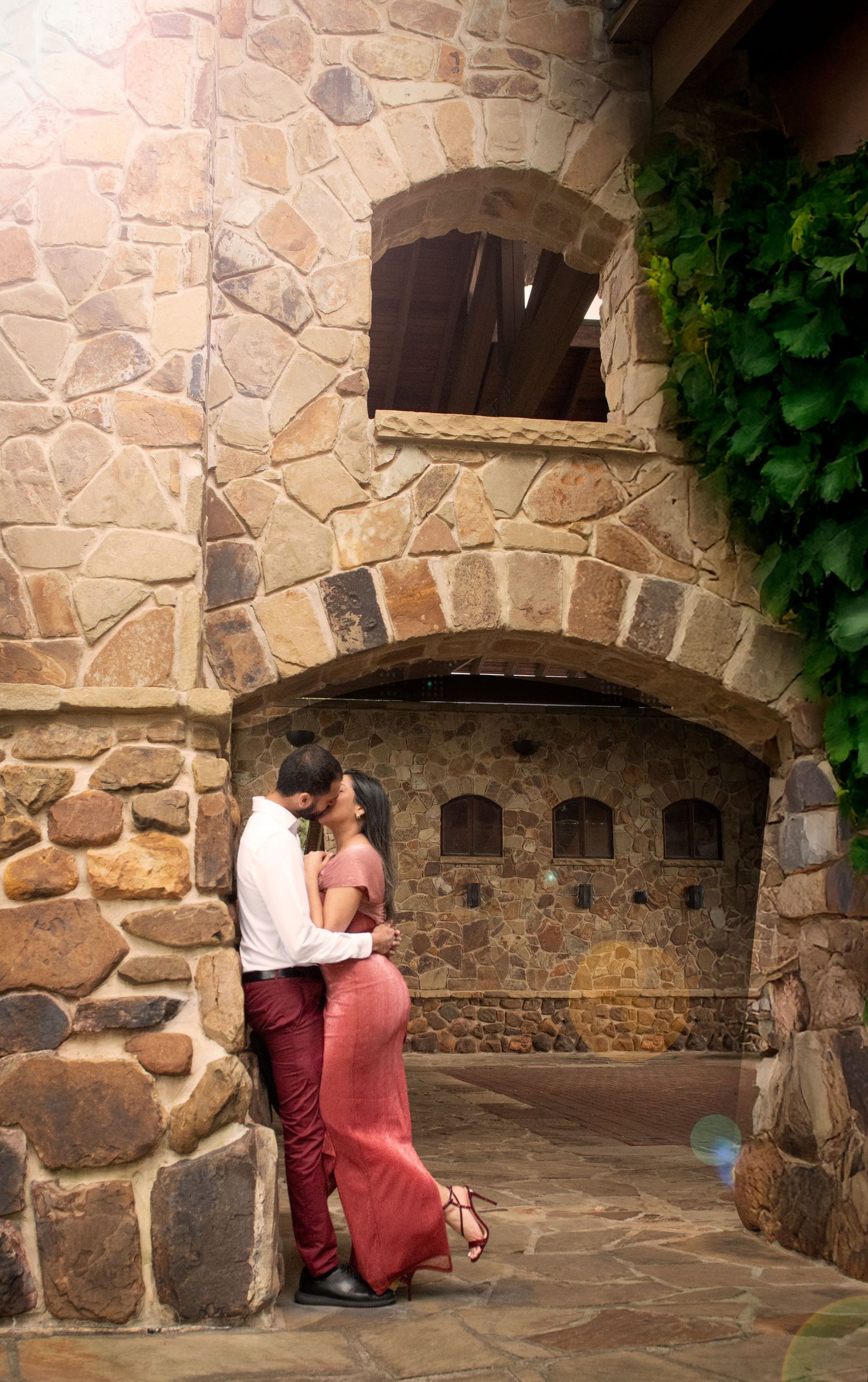 Romantic couple embracing against a rustic stone wall arch during an outdoor engagement photo session.