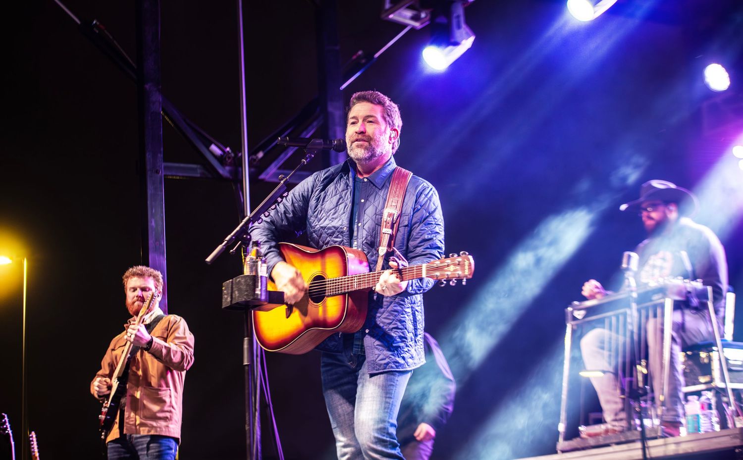 Male country musician plays acoustic guitar on stage under blue spotlights at an outdoor concert.