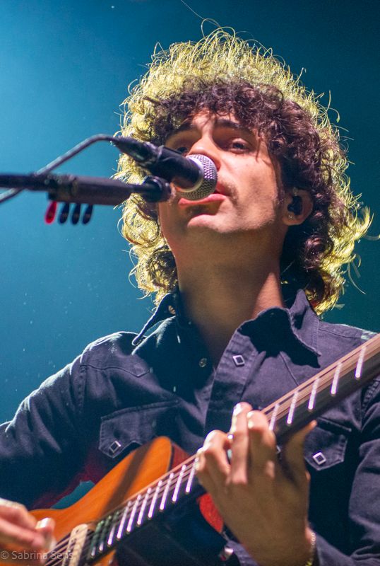 Young male musician with curly hair playing acoustic guitar and singing into microphone on stage.