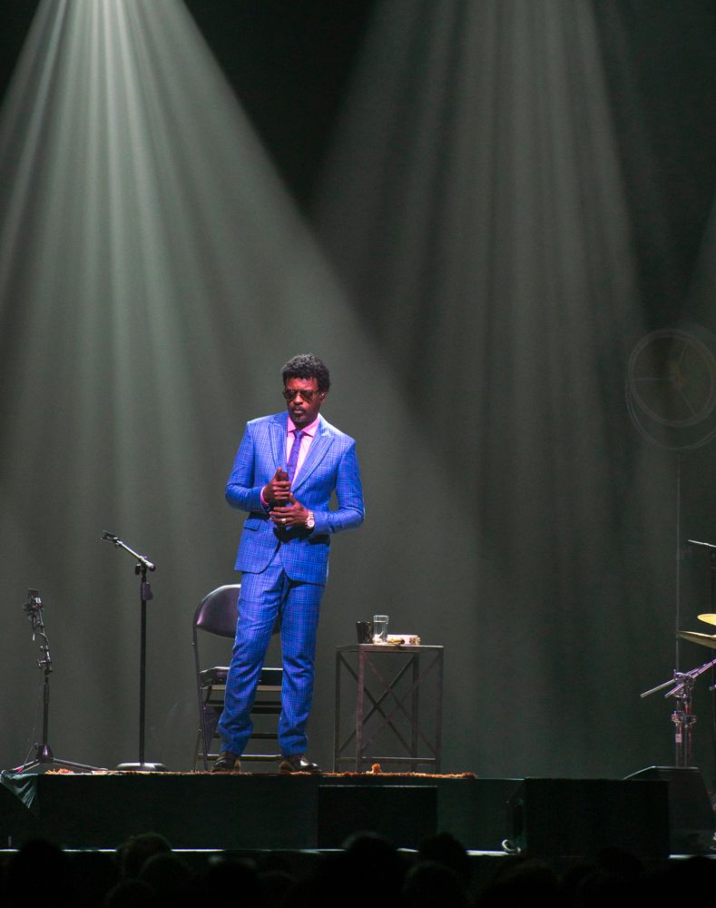Male vocalist in bright blue suit performing solo on a dramatic spotlight-lit concert stage.