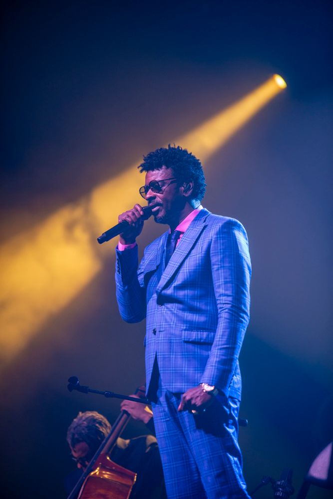 Male performer in blue plaid suit singing into microphone on stage under dramatic yellow and blue spotlights.