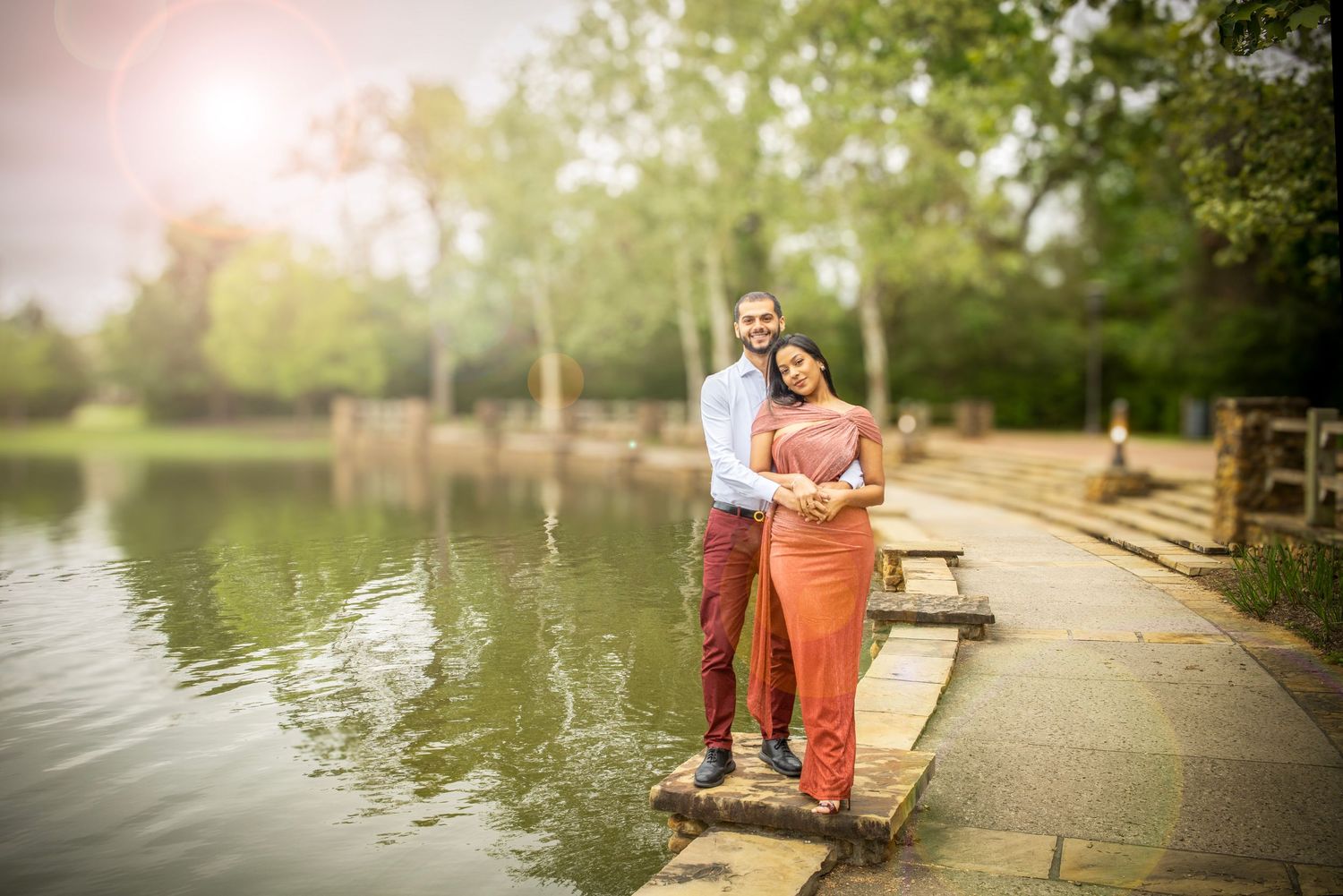 Couple embracing on a stone pathway beside a serene lake surrounded by lush trees.