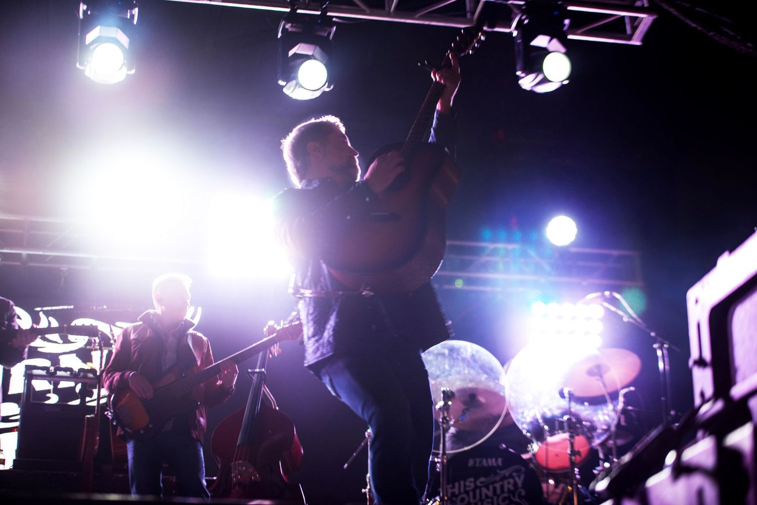 A rock band performs energetically on a dark stage with dramatic overhead spotlights and a visible drum kit.