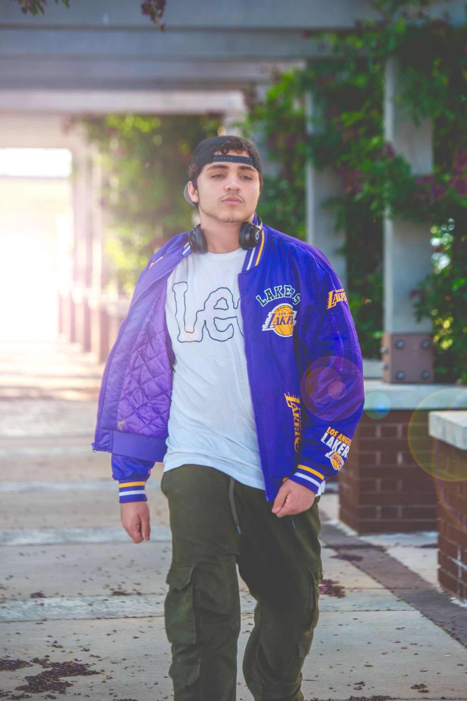 Young man in purple LA Lakers jacket and cap walking under sunlit outdoor corridor with ivy-covered columns.